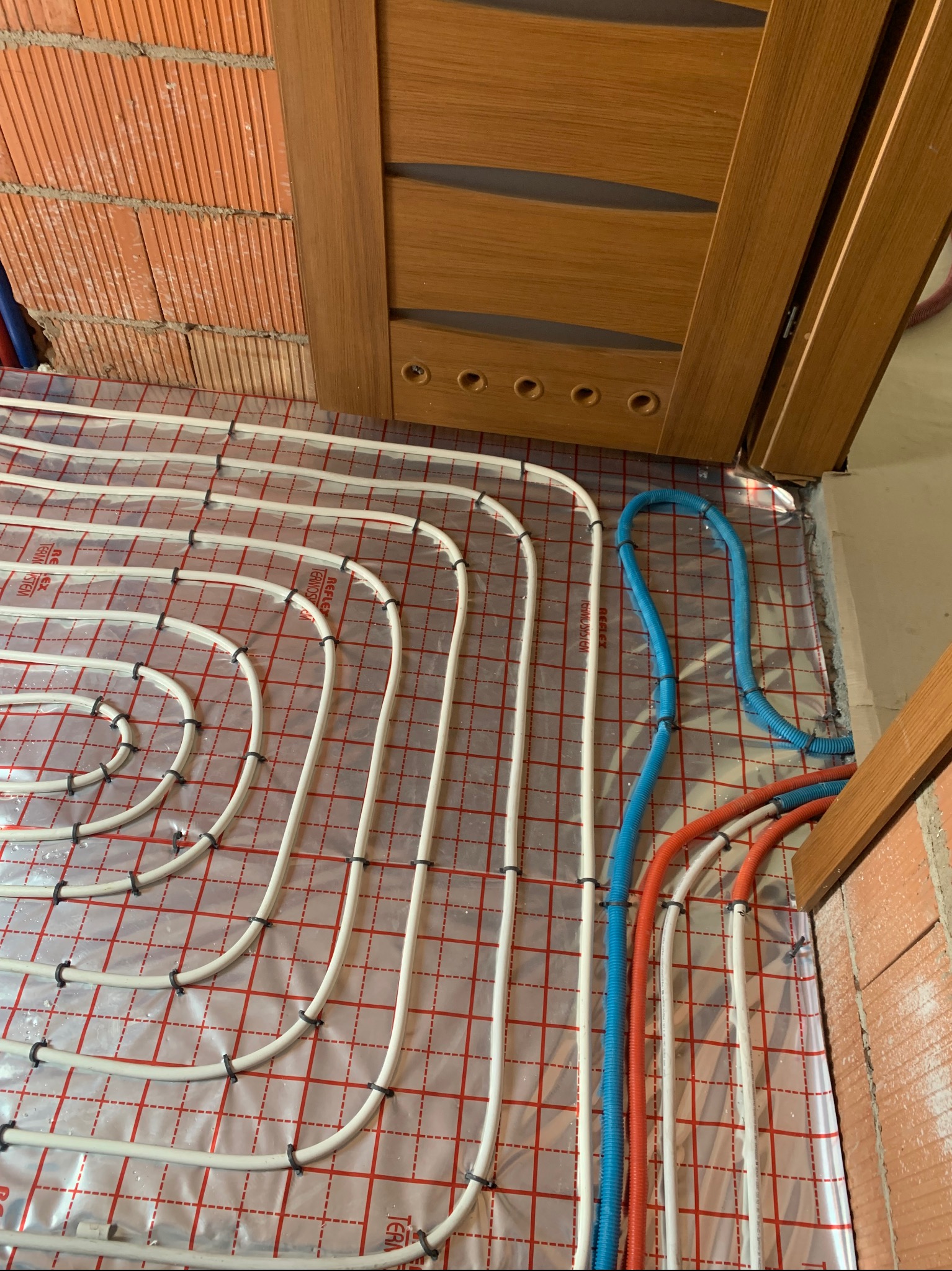 Białe rurki ogrzewania podłogowego ułożone na folii z czerwoną kratką, przymocowane czarnymi uchwytami. Widoczne fragmenty ściany z cegły i drewnianej ościeżnicy.