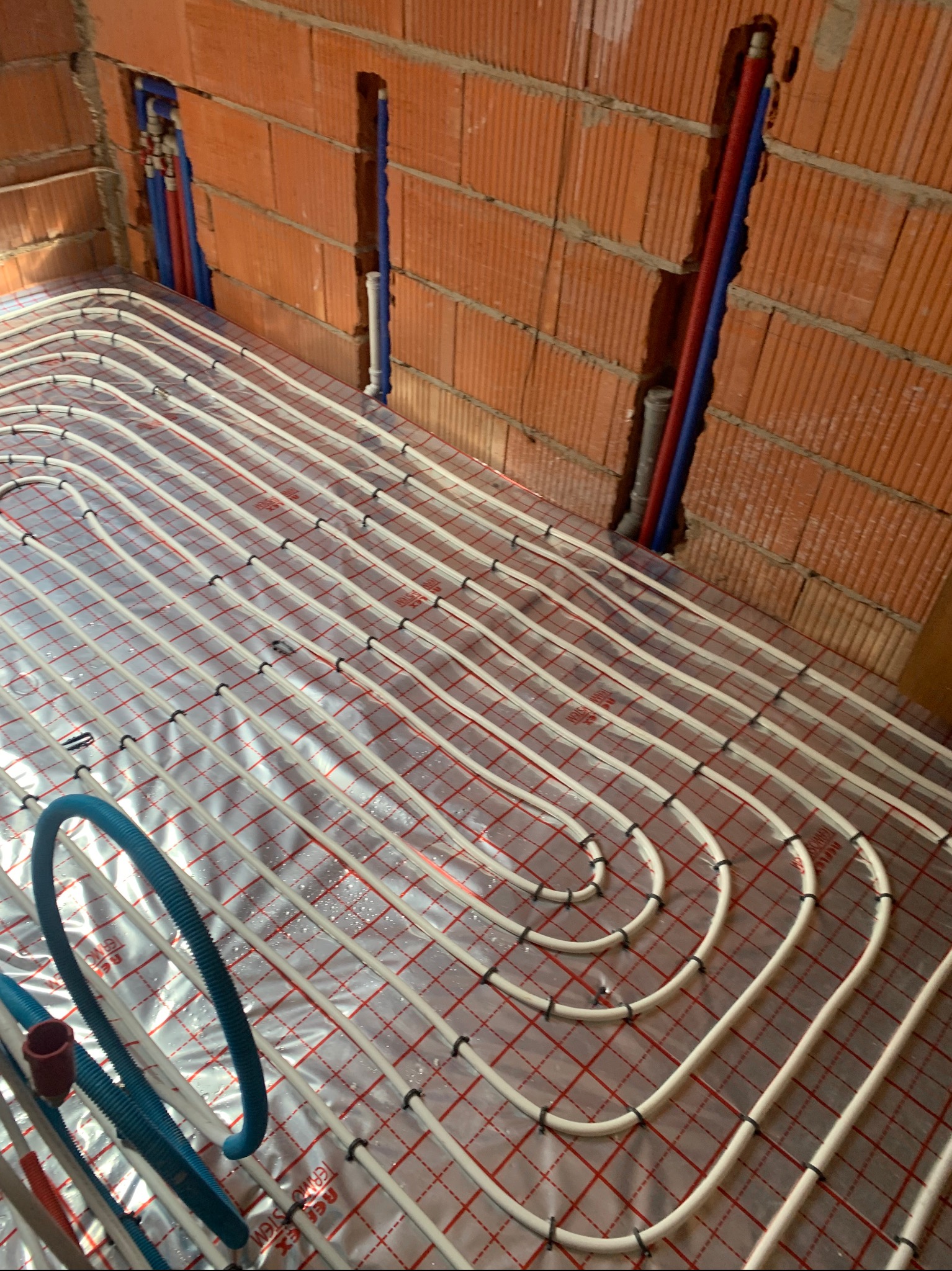 Białe rury ogrzewania podłogowego ułożone na folii z czerwoną siatką, przymocowane czarnymi uchwytami. W tle ściana z czerwonej cegły z pionowymi rurami.