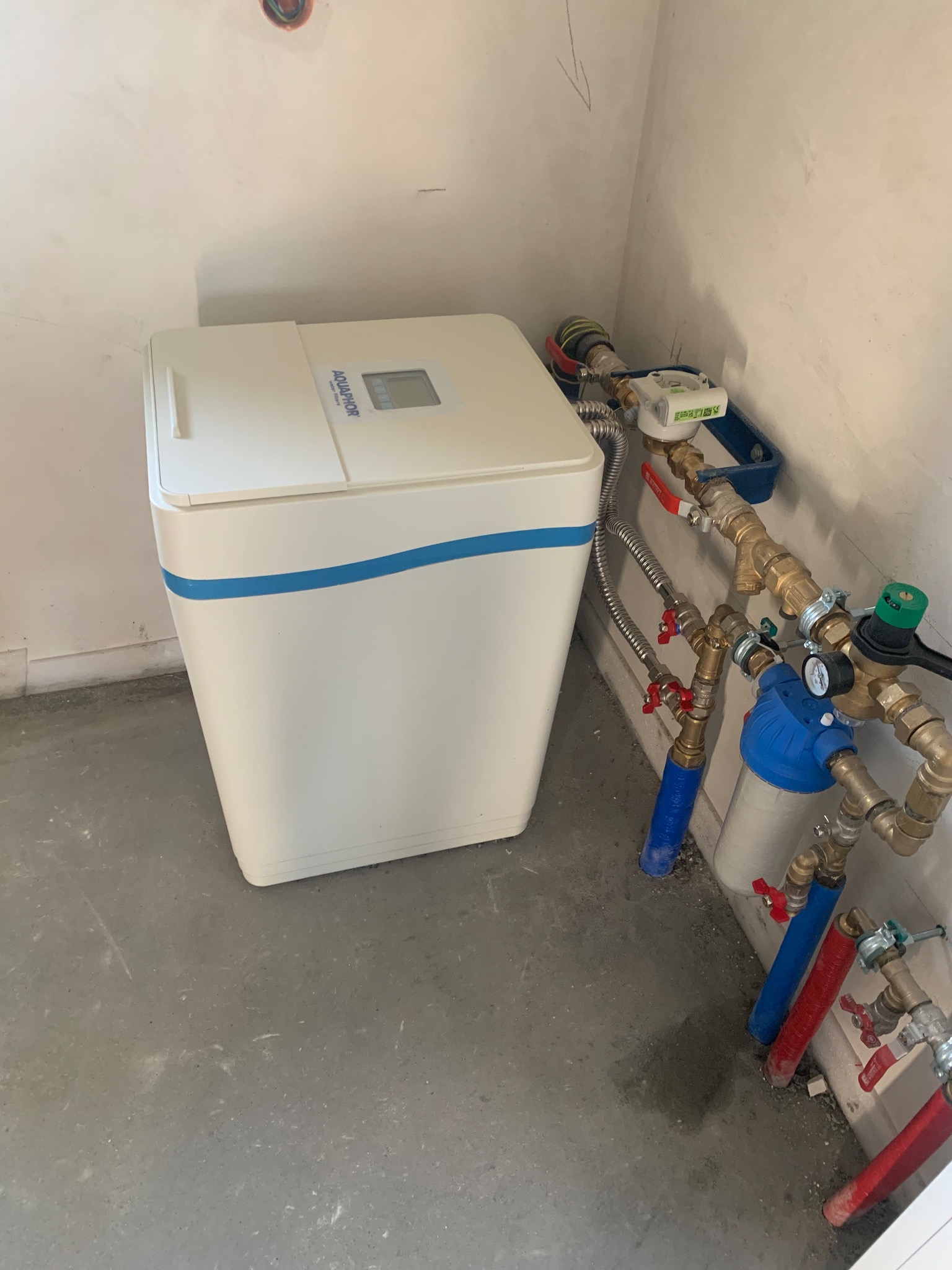Biały zmiękczacz wody Aquaphor z niebieskim paskiem, podłączony do instalacji z zaworami i filtrem, na szarym betonowym podłożu w surowym wnętrzu.