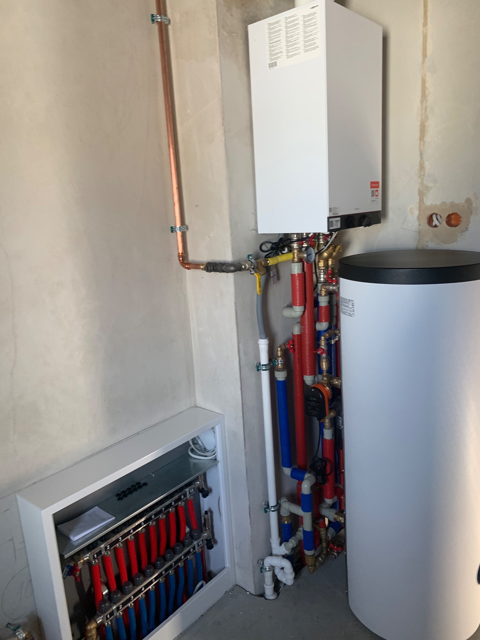 Biały piec grzewczy z zbiornikiem i rozdzielaczem ogrzewania podłogowego, połączone rurami miedzianymi i plastikowymi w surowym pomieszczeniu.