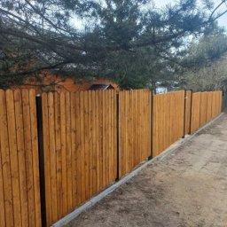 Dawid Ogrodzenia - Długie, drewniane ogrodzenie sztachetowe w ciepłym odcieniu brązu, z czarnymi słupkami, otoczone ziemią i zielenią. Widok z perspektywy gruntu.