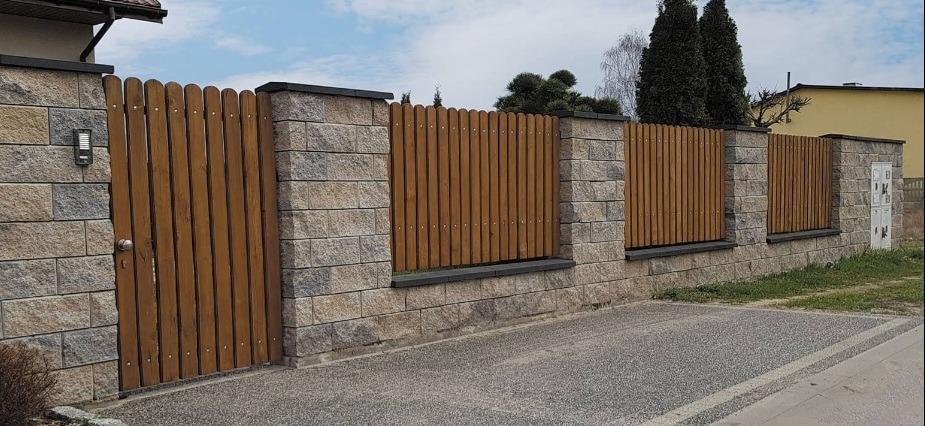 Drewniana brama i ogrodzenie z elementami betonowymi, z widocznym oświetleniem przy bramie, na tle błękitnego nieba i zieleni.