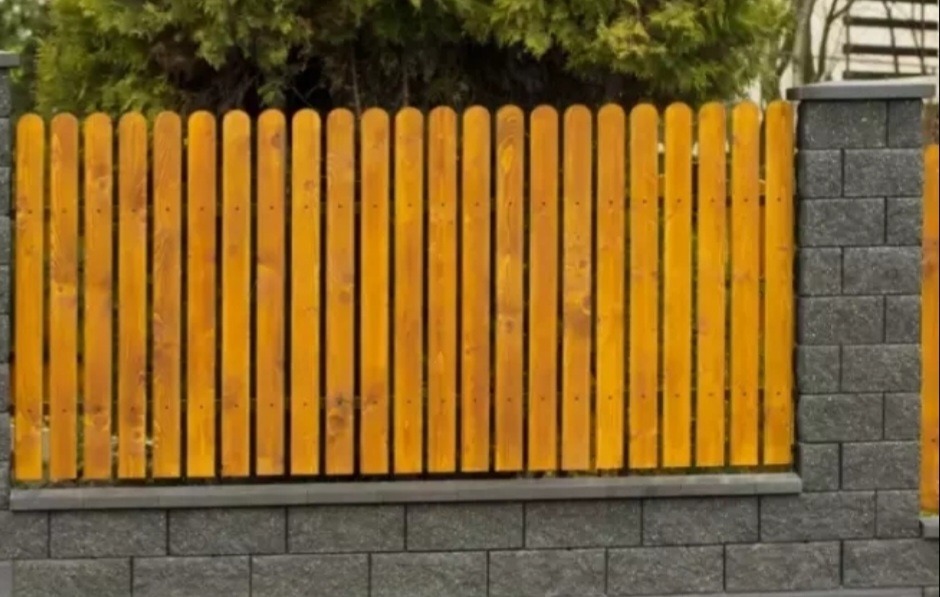 Drewniane sztachety ogrodzenia na podmurówce z szarej kostki, Białystok. Jasne drewno kontrastuje z ciemną kostką, a zieleń w tle dodaje naturalności.