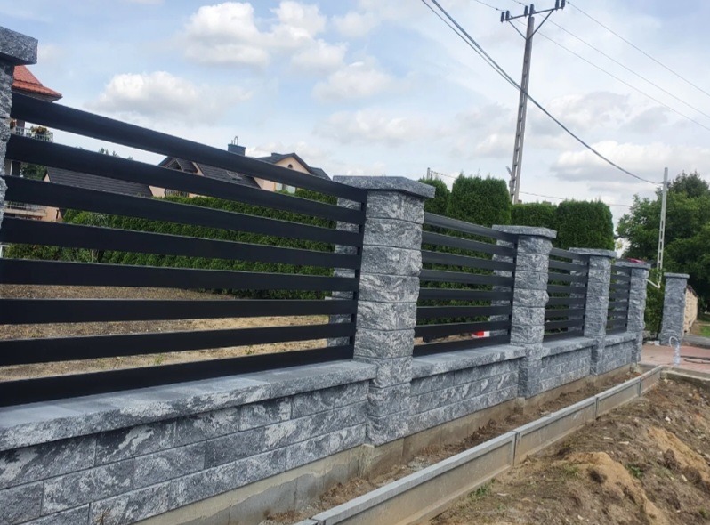 Nowoczesne ogrodzenie z szarego kamienia i czarnych, poziomych paneli. Widok z boku, z fragmentem domu w tle i słupem elektrycznym po prawej stronie.