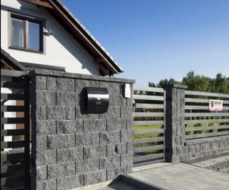 Nowoczesne ogrodzenie z szarego kamienia łupanego i metalowych przęseł, z czarną skrzynką na listy, na tle domu z oknem i błękitnego nieba.