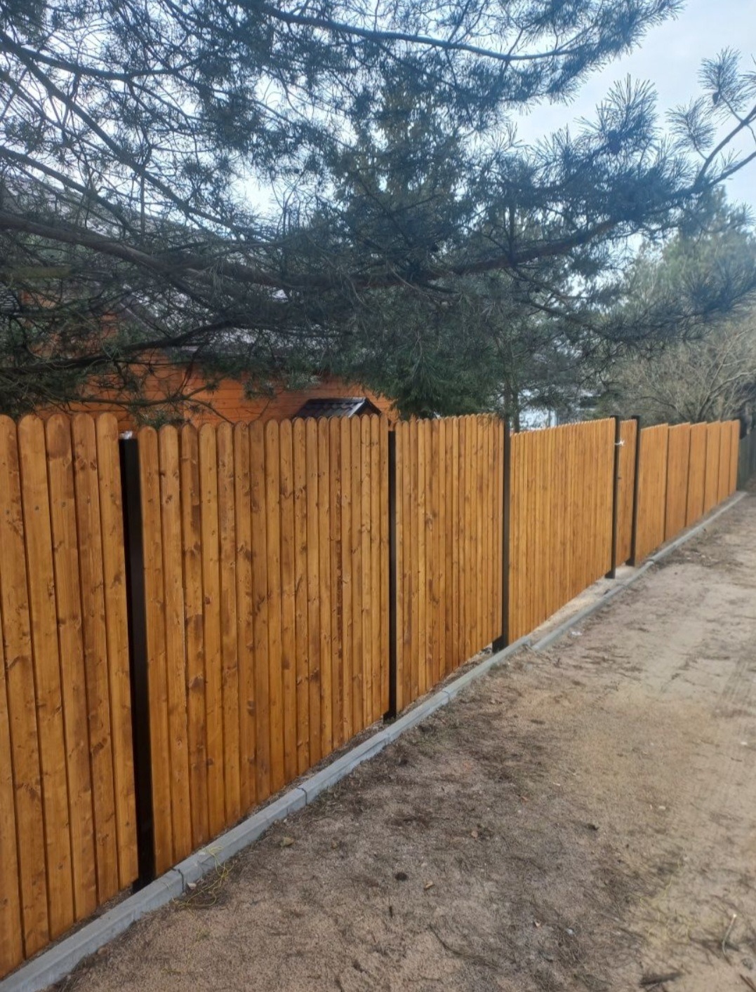 Długie, drewniane ogrodzenie sztachetowe w ciepłym odcieniu brązu, z czarnymi słupkami, otoczone ziemią i zielenią. Widok z perspektywy gruntu.