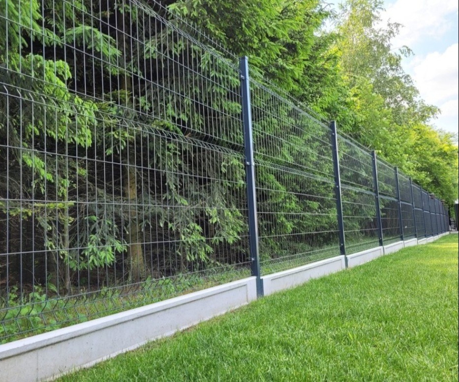 Nowoczesne ogrodzenie panelowe w kolorze grafitowym, osadzone na betonowej podmurówce, otoczone zielonym trawnikiem i gęstą roślinnością.