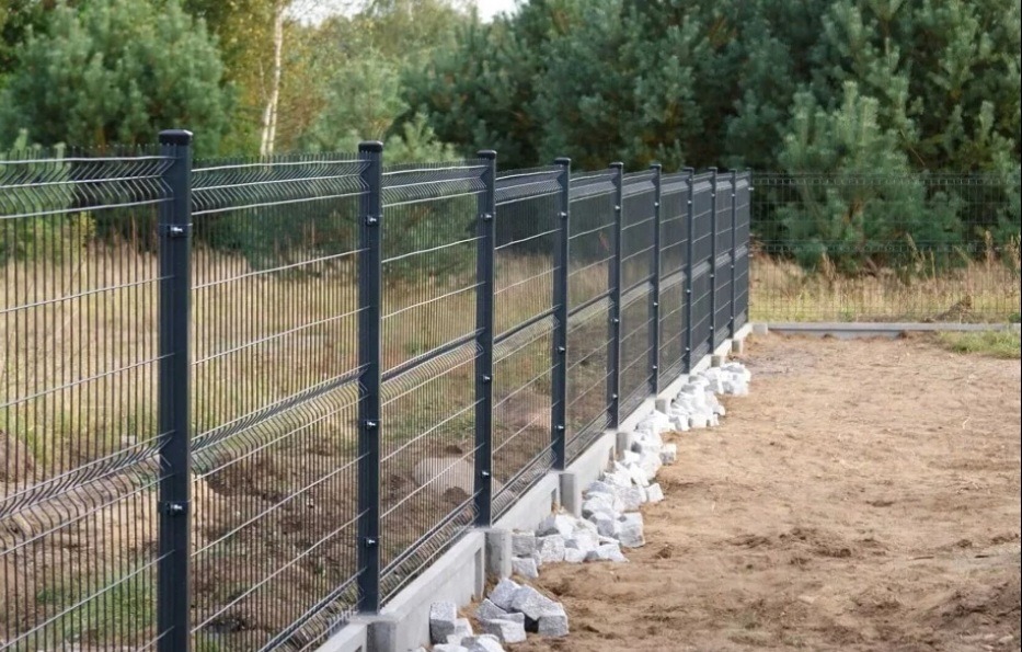 Nowoczesne ogrodzenie panelowe w kolorze grafitowym, osadzone na betonowej podmurówce z kamieniami u podstawy, na tle zieleni drzew. Widok z boku.