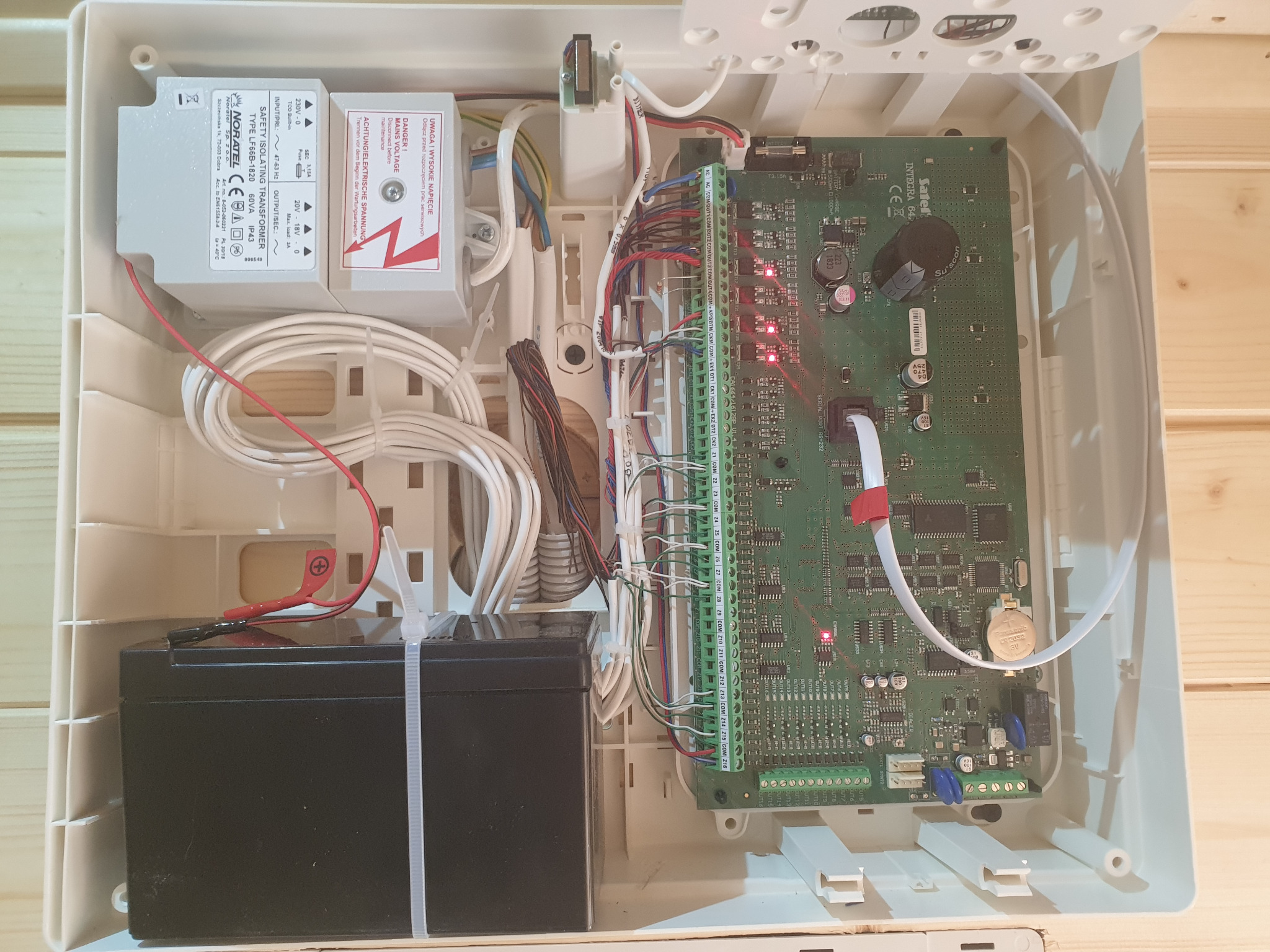 Otwarta obudowa centrali alarmowej z widoczną elektroniką, akumulatorem i transformatorem. Widoczne liczne przewody i złącza. Elementy systemu alarmowego wewnątrz białej skrzynki.