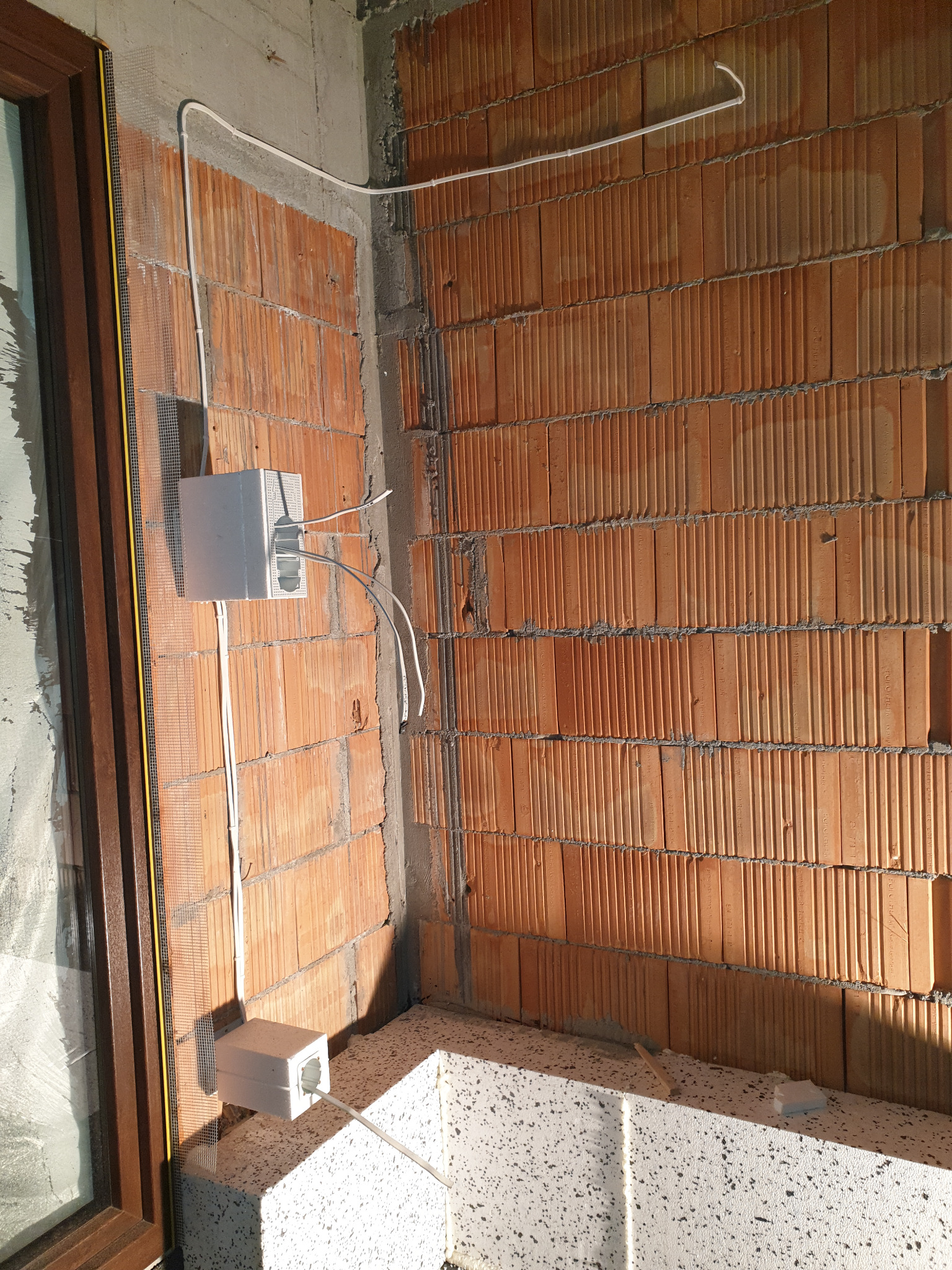 Wewnętrzna instalacja elektryczna w trakcie montażu: puszki podtynkowe z przewodami na ceglanej ścianie, widoczne okno z brązową ramą i fragment ściany z betonu komórkowego.