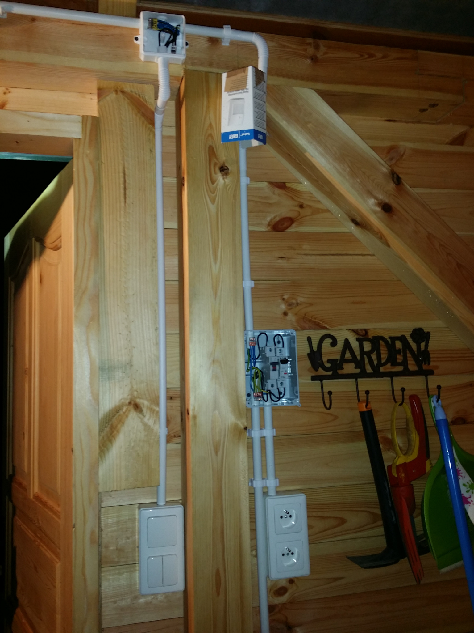 Instalacja elektryczna w drewnianym domku: białe kable w peszlach, puszki, gniazdka i włączniki na ścianie z desek. Widoczne narzędzia ogrodnicze.