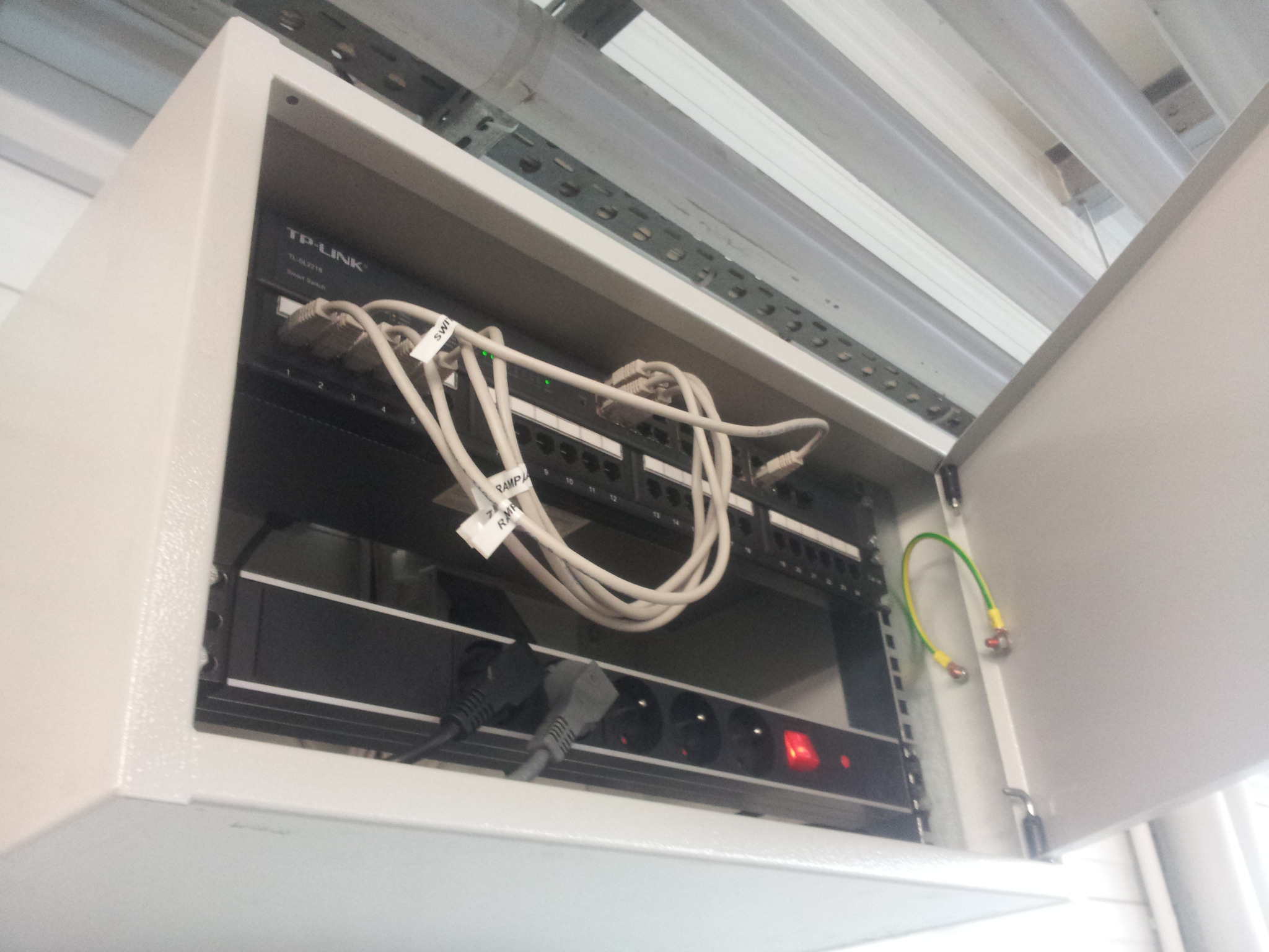 Otwarta szafa rack z patch panelem, switchem TP-Link i listwą zasilającą. Widoczne kable sieciowe z etykietami i przewód uziemiający na drzwiach szafy.