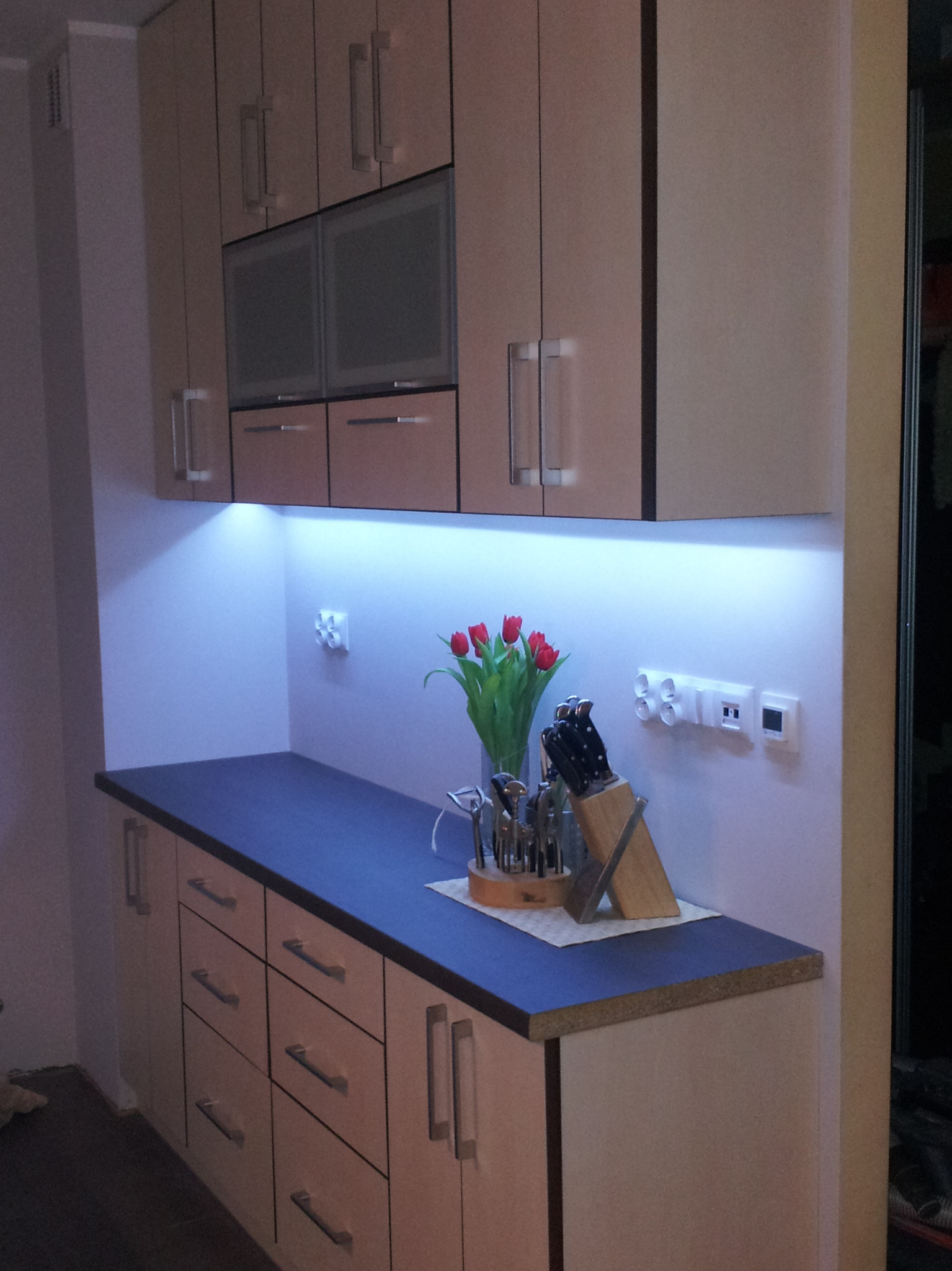 Nowoczesna kuchnia z podświetleniem LED podszafkowym, gniazdkami i włącznikami na ścianie. Na blacie wazon z czerwonymi tulipanami i stojak z nożami.