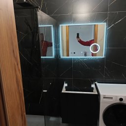 Jan Wróblewski - Nowoczesna łazienka z czarnymi płytkami imitującymi marmur, podświetlanym lustrem i kabiną prysznicową typu walk-in. Widoczna pralka i częściowo zamontowana szafka pod umywalkę.