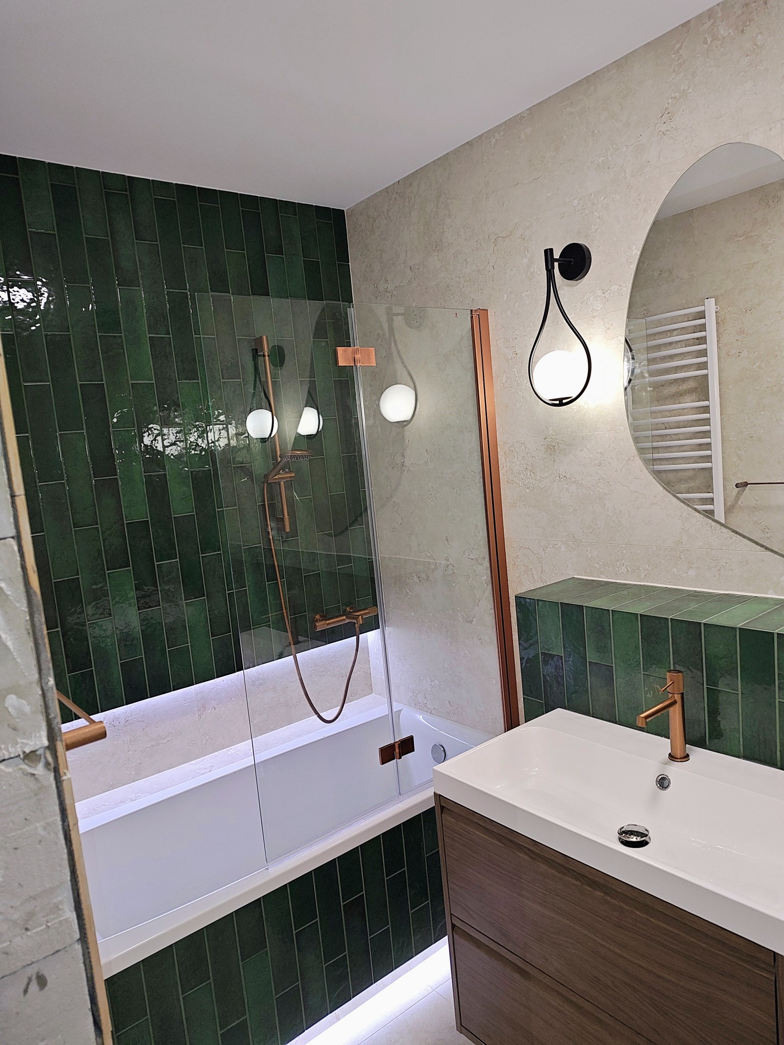 Nowoczesna łazienka z zielonymi, błyszczącymi płytkami typu cegiełka, armaturą w kolorze miedzi i owalnym lustrem. Widoczna wanna z parawanem i umywalka z szafką.