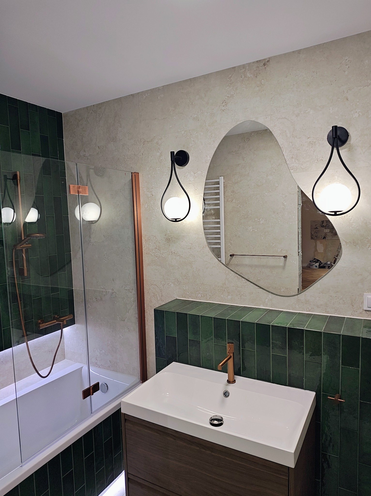 Nowoczesna łazienka z zielonymi płytkami, asymetrycznym lustrem i designerskimi lampami. Umywalka z szafką w kolorze drewna. Kabina prysznicowa z elementami miedzianymi.