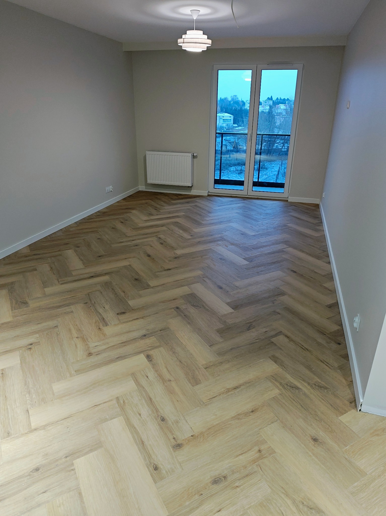 Jasny parkiet ułożony w jodełkę w pustym pokoju z balkonem. Widoczne białe listwy przypodłogowe, grzejnik i lampa sufitowa. Za oknem widok na zabudowę miejską.
