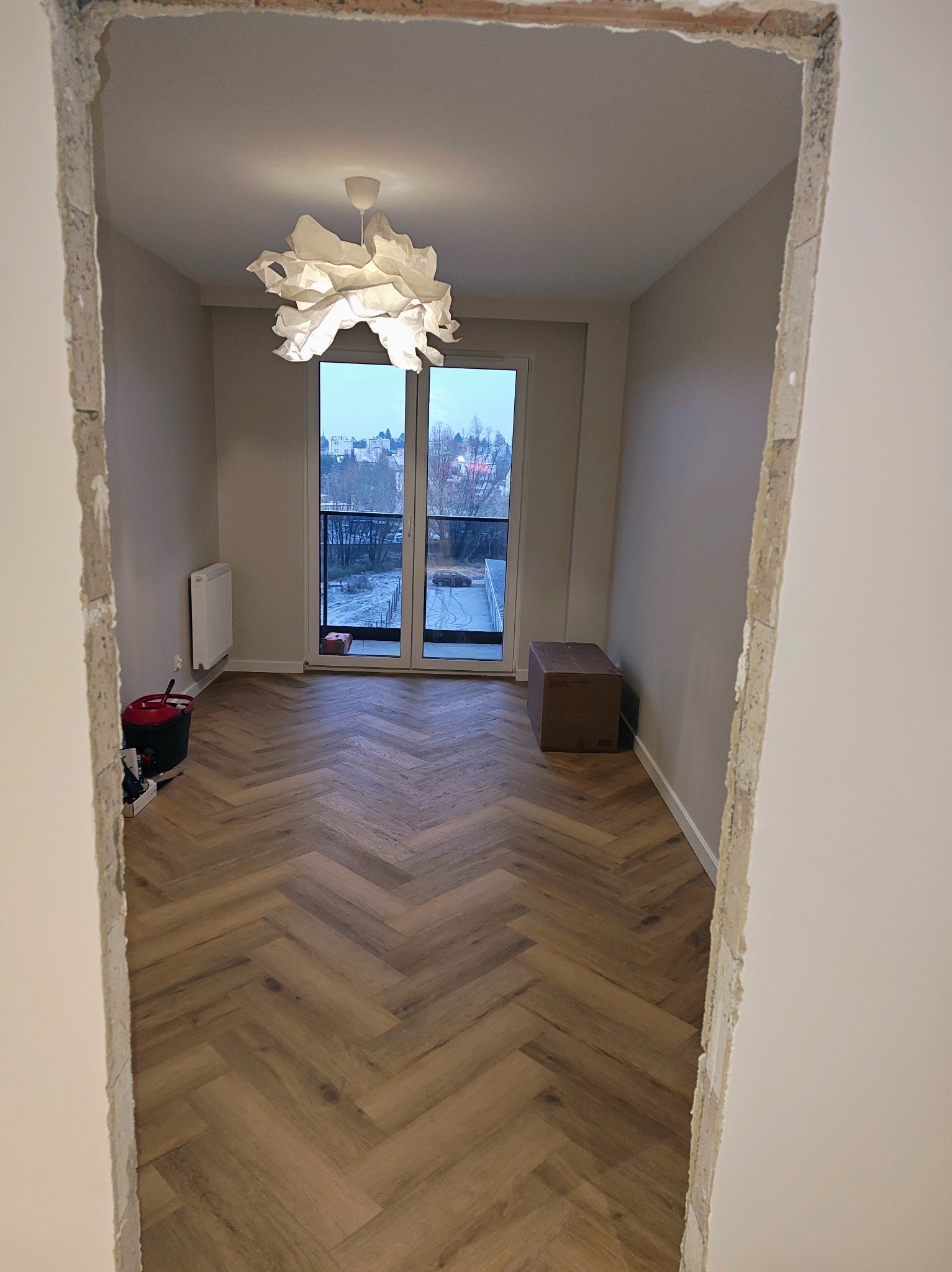 Pokój z ułożonym parkietem w jodełkę, widok przez surowy otwór drzwiowy. Nowoczesna lampa, balkon z widokiem na miasto w tle. Etap prac wykończeniowych.