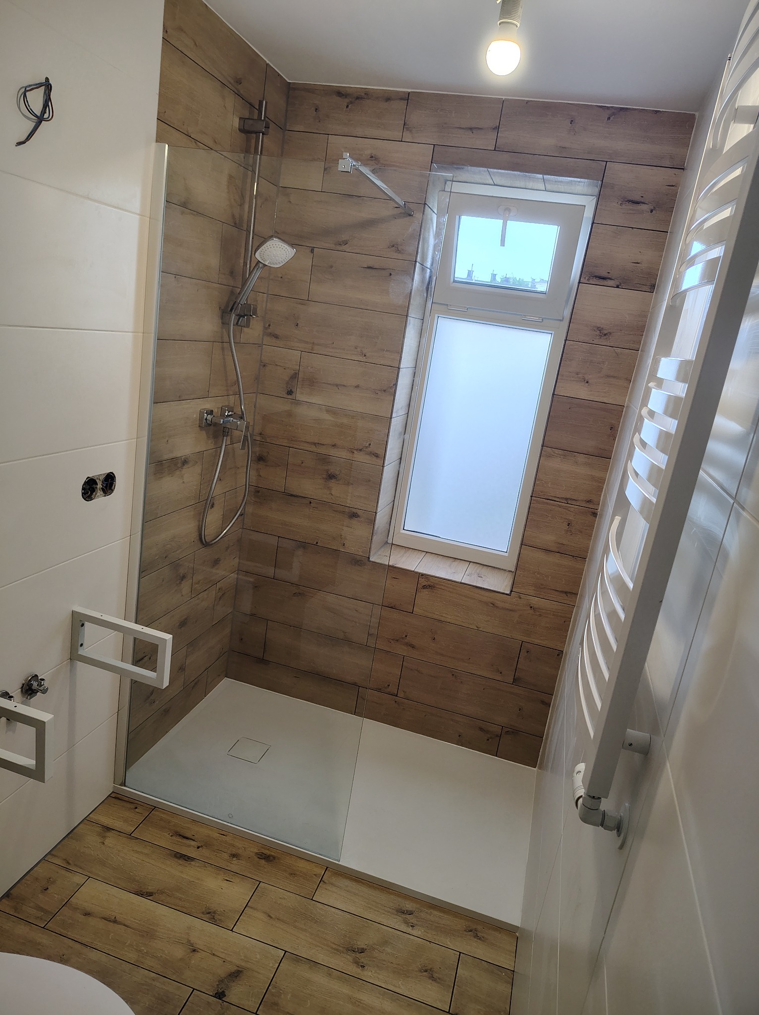 Nowoczesna kabina prysznicowa z drewnopodobnymi płytkami, białym brodzikiem i grzejnikiem drabinkowym. Widoczne okno w skosie i niedokończone podłączenie elektryczne.