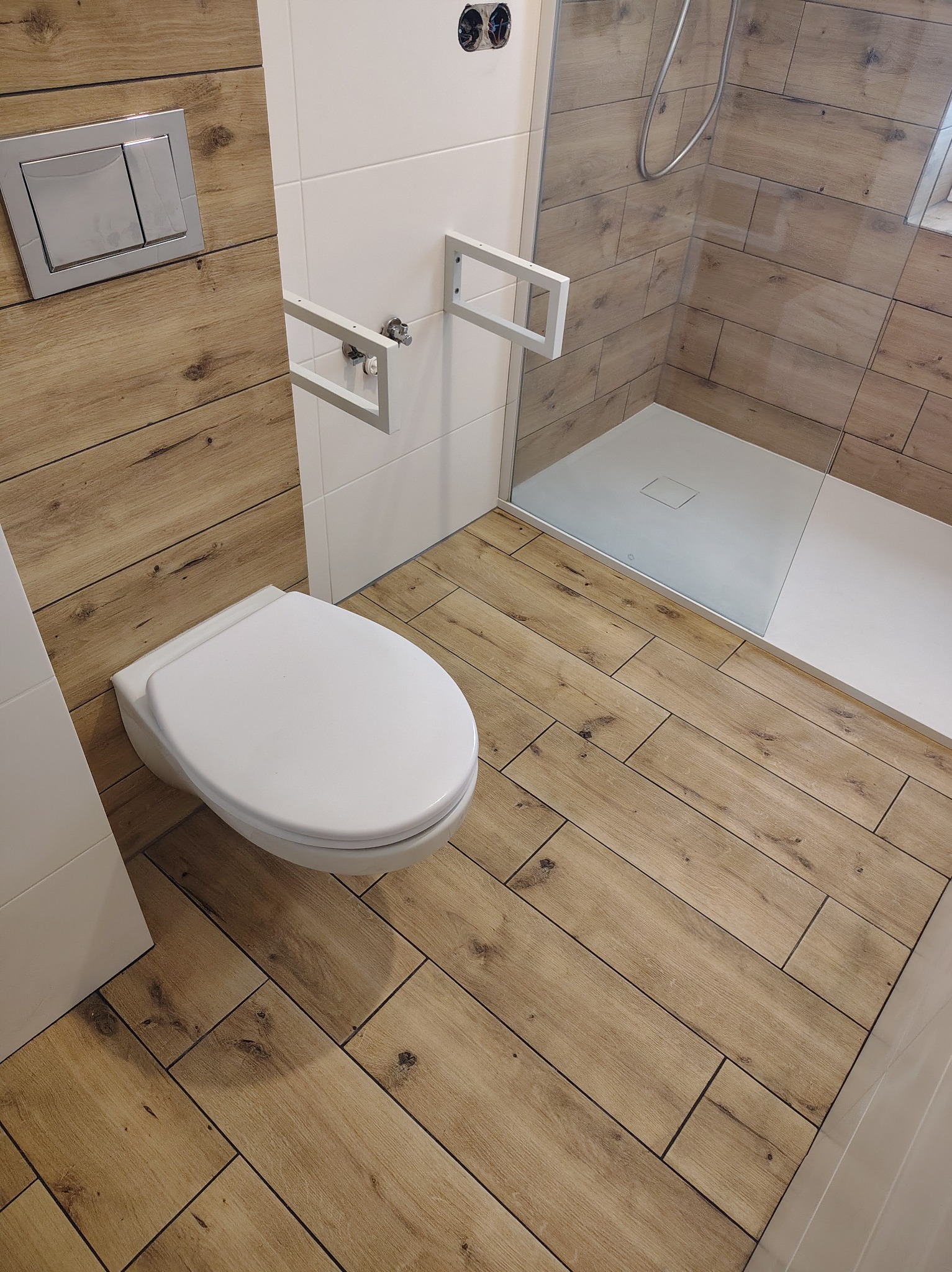Nowoczesna łazienka z drewnopodobnymi płytkami, kabiną prysznicową i podwieszaną toaletą. Widoczne białe stelaże pod umywalkę i przycisk spłukujący.