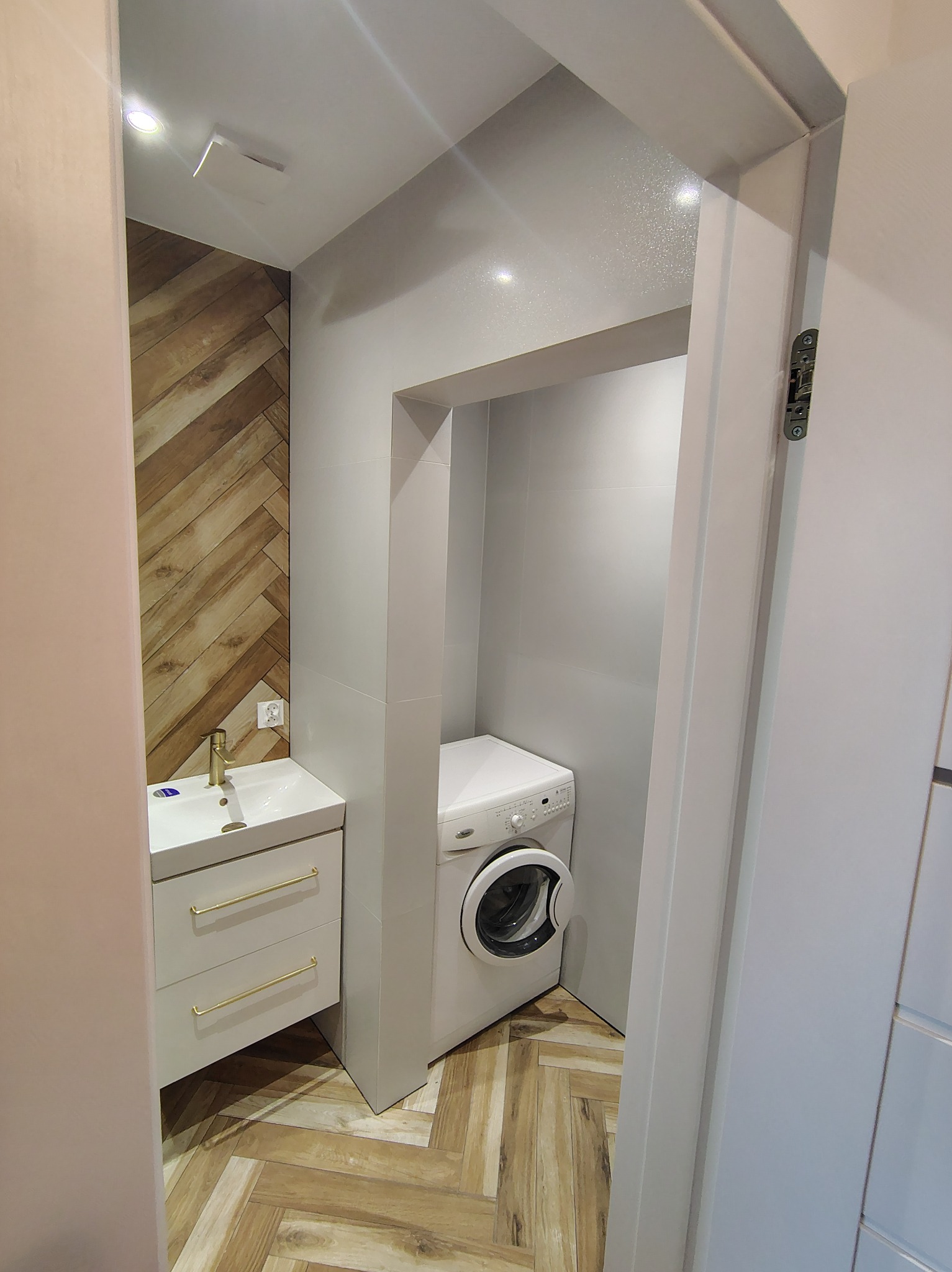 Nowoczesna łazienka po remoncie: biała umywalka z szafką, pralka w zabudowie, drewnopodobne płytki ułożone w jodełkę na ścianie i podłodze, widok zza drzwi.