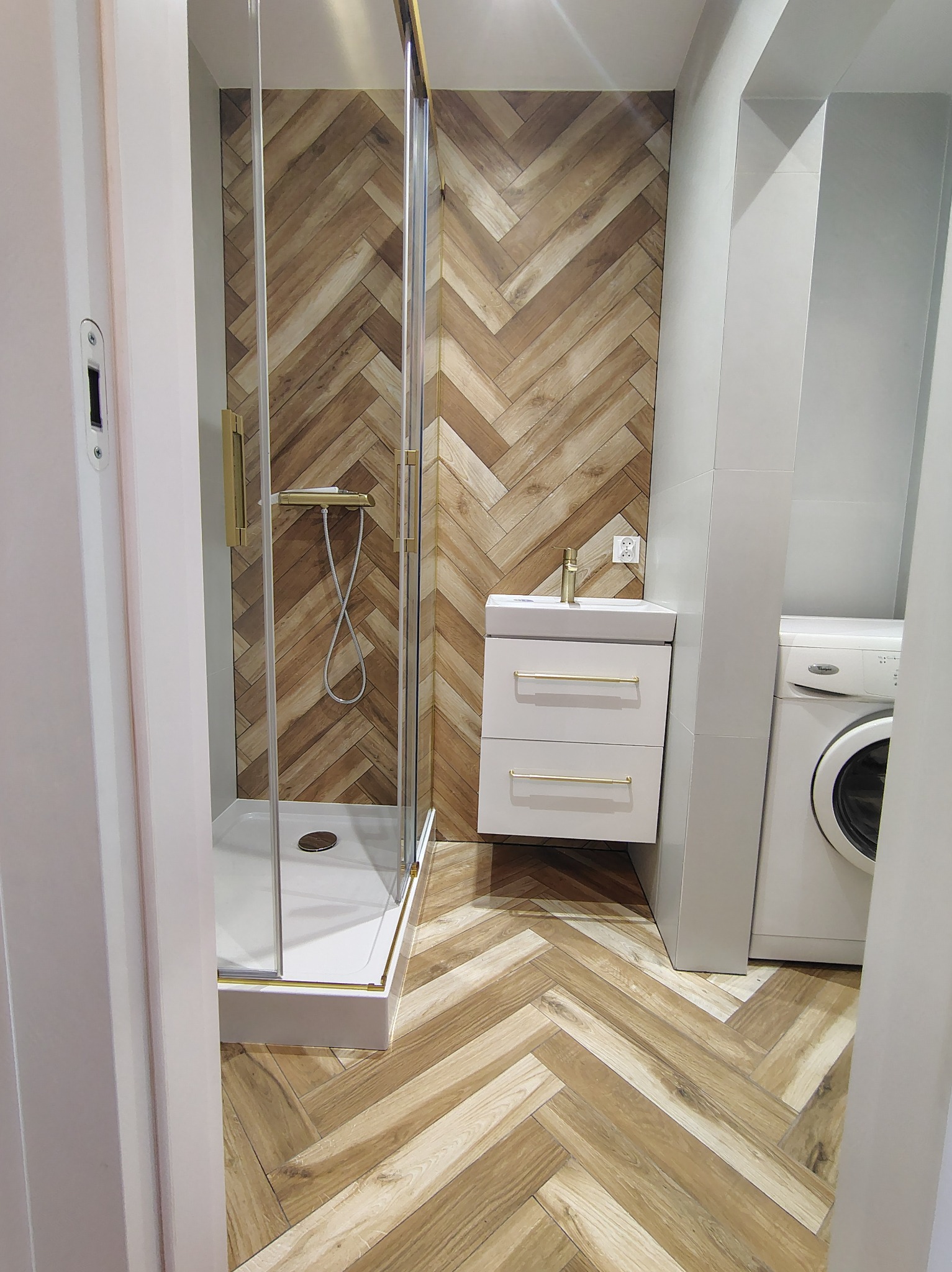 Nowoczesna łazienka z kabiną prysznicową, umywalką z szafką i pralką. Parkiet ułożony w jodełkę, złote akcenty. Widok zza futryny.