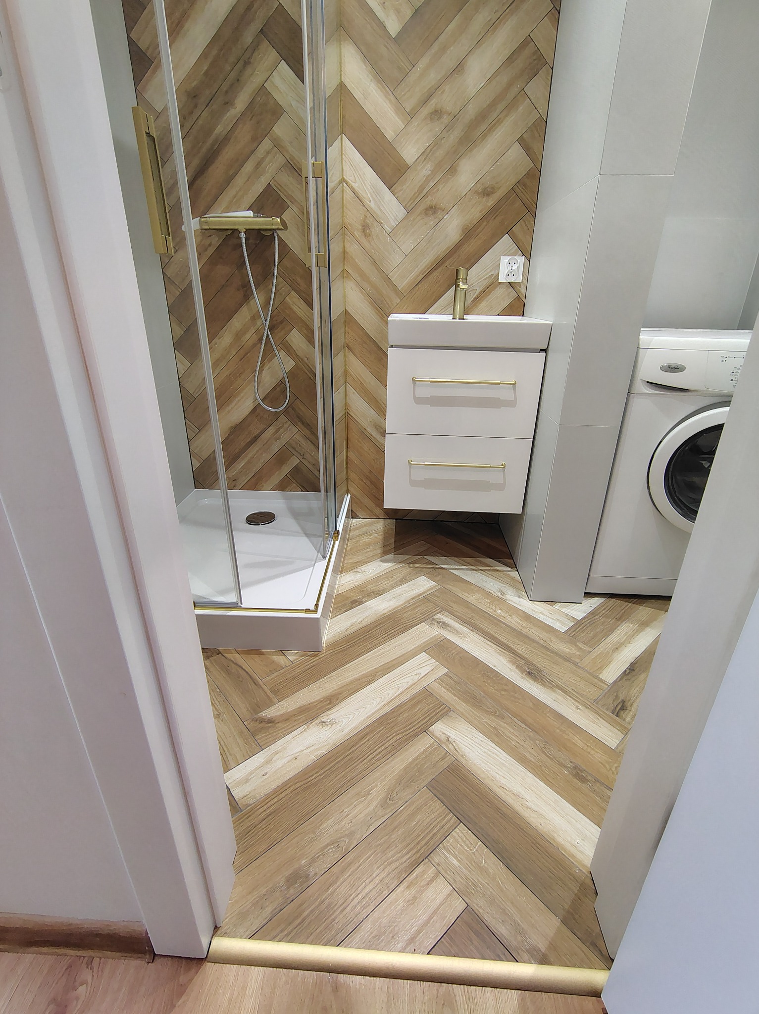 Nowoczesna łazienka z kabiną prysznicową, umywalką z szafką i pralką. Parkiet ułożony w jodełkę, złote akcenty. Widok zza framugi drzwi.