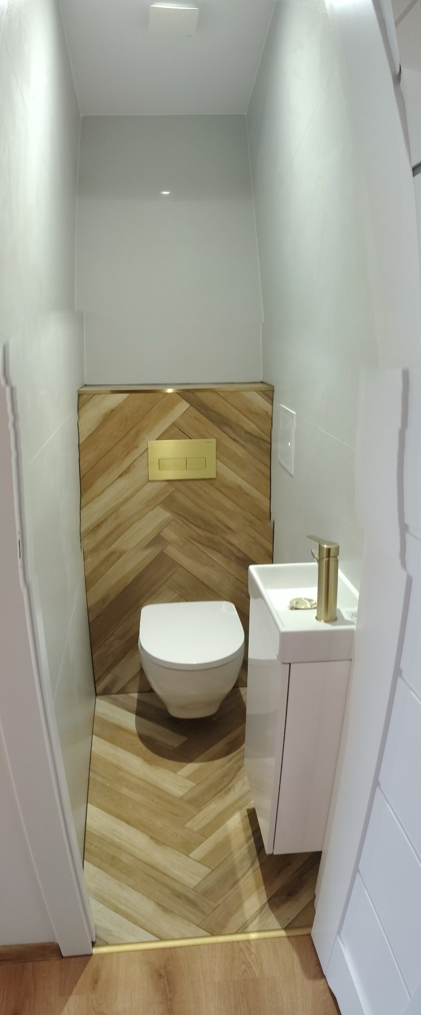 Nowoczesna toaleta z drewnopodobnymi płytkami ułożonymi w jodełkę na podłodze i ścianie za toaletą. Biała umywalka z złotą baterią. Minimalistyczny design.