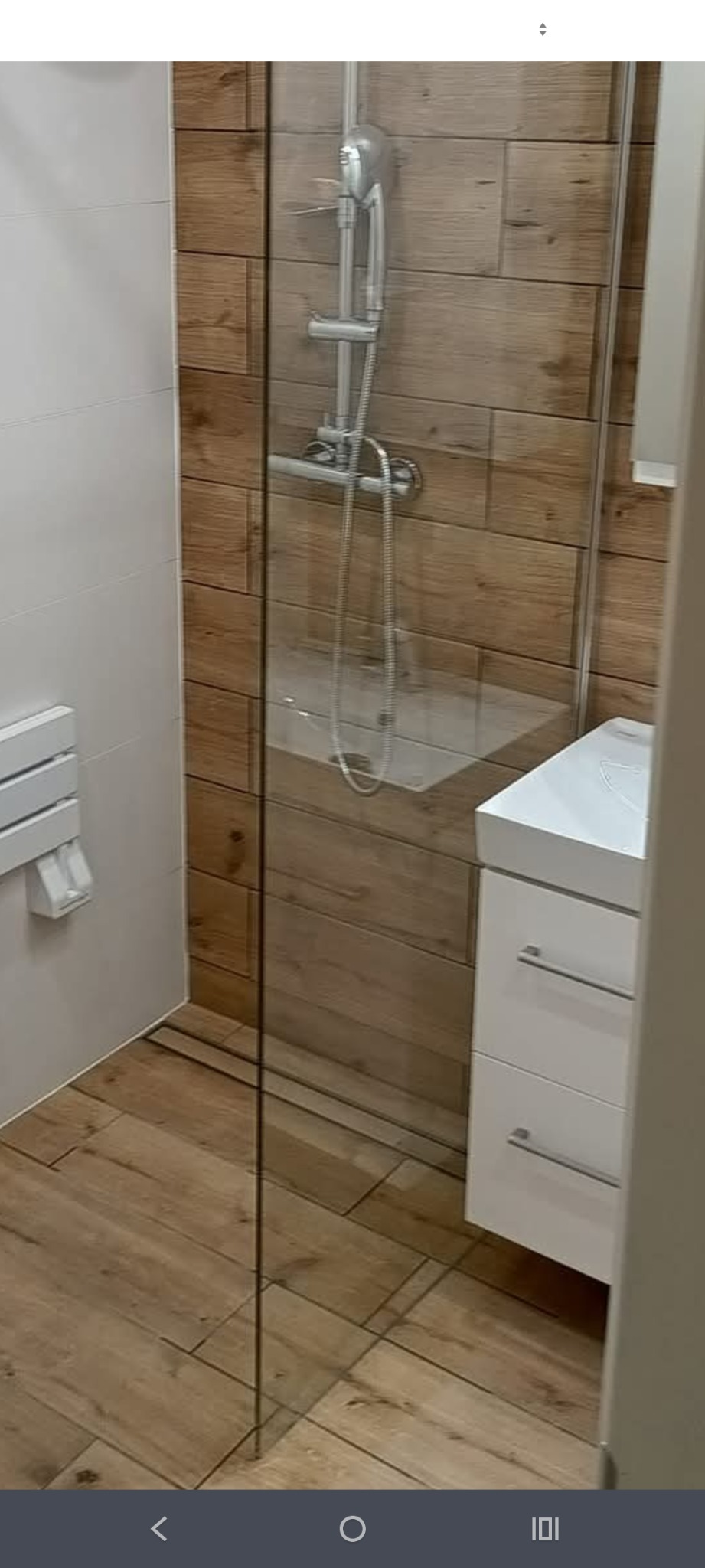 Nowoczesna łazienka z kabiną prysznicową typu walk-in, wykończona płytkami imitującymi drewno, z białą szafką pod umywalką i grzejnikiem drabinkowym.