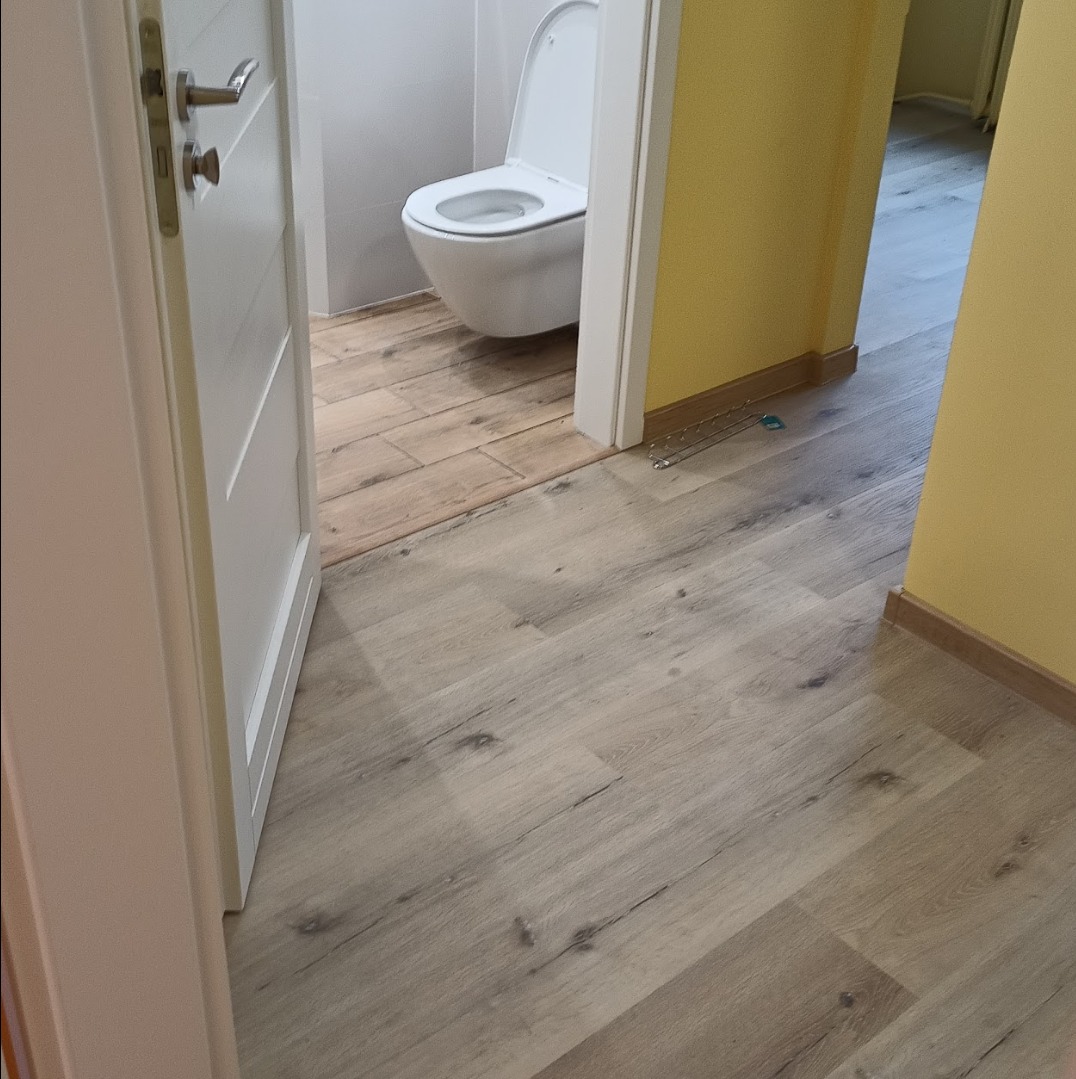 Otwarta biała łazienkowa z toaletą i podłogą z paneli imitujących drewno, widok z korytarza z żółtymi ścianami. Na podłodze leżą metalowe akcesoria.