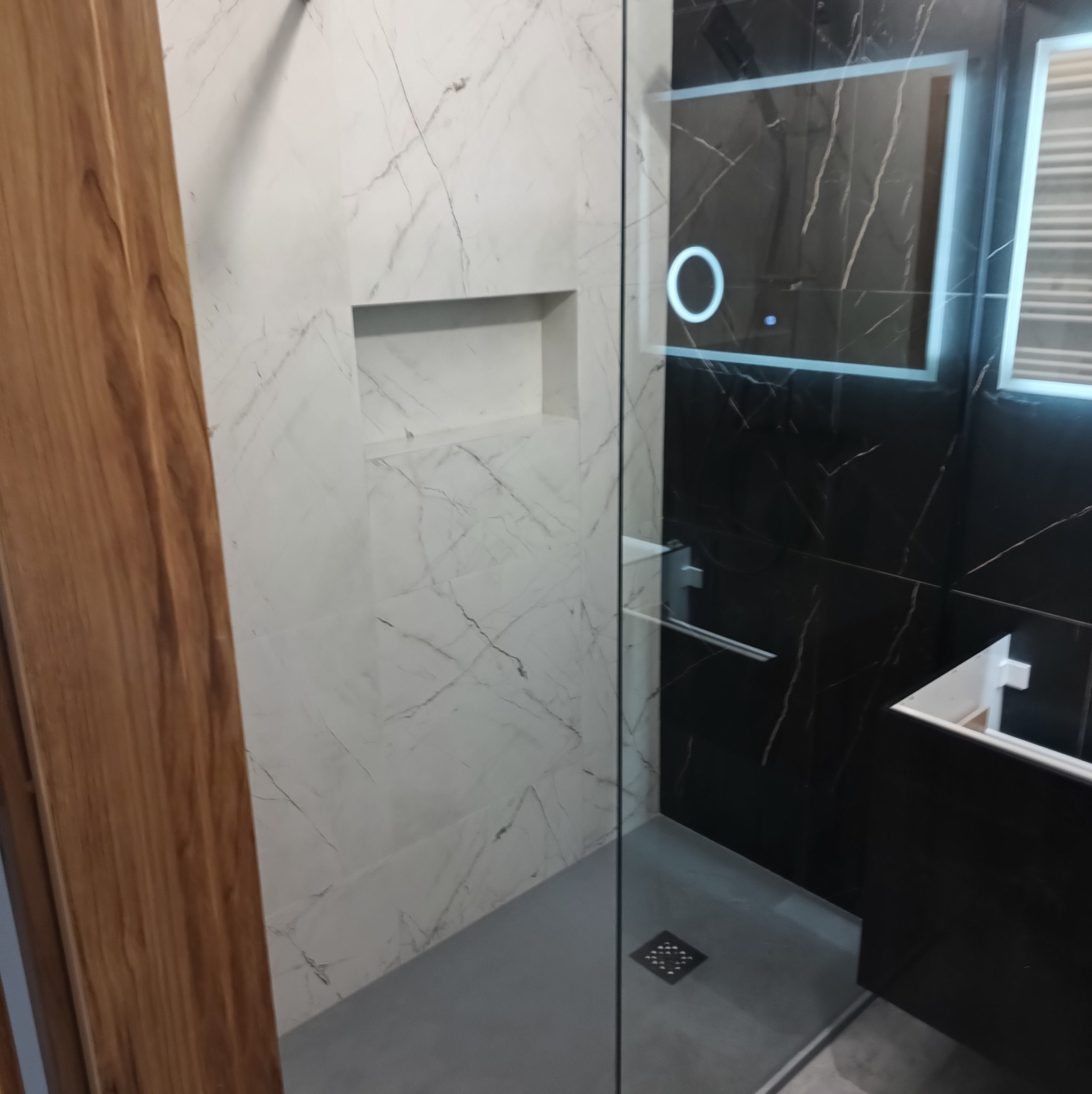 Nowoczesna kabina prysznicowa z czarno-białymi płytkami imitującymi marmur, podświetlanym lustrem i wnęką w ścianie. Minimalistyczny design, szklana ściana kabiny.