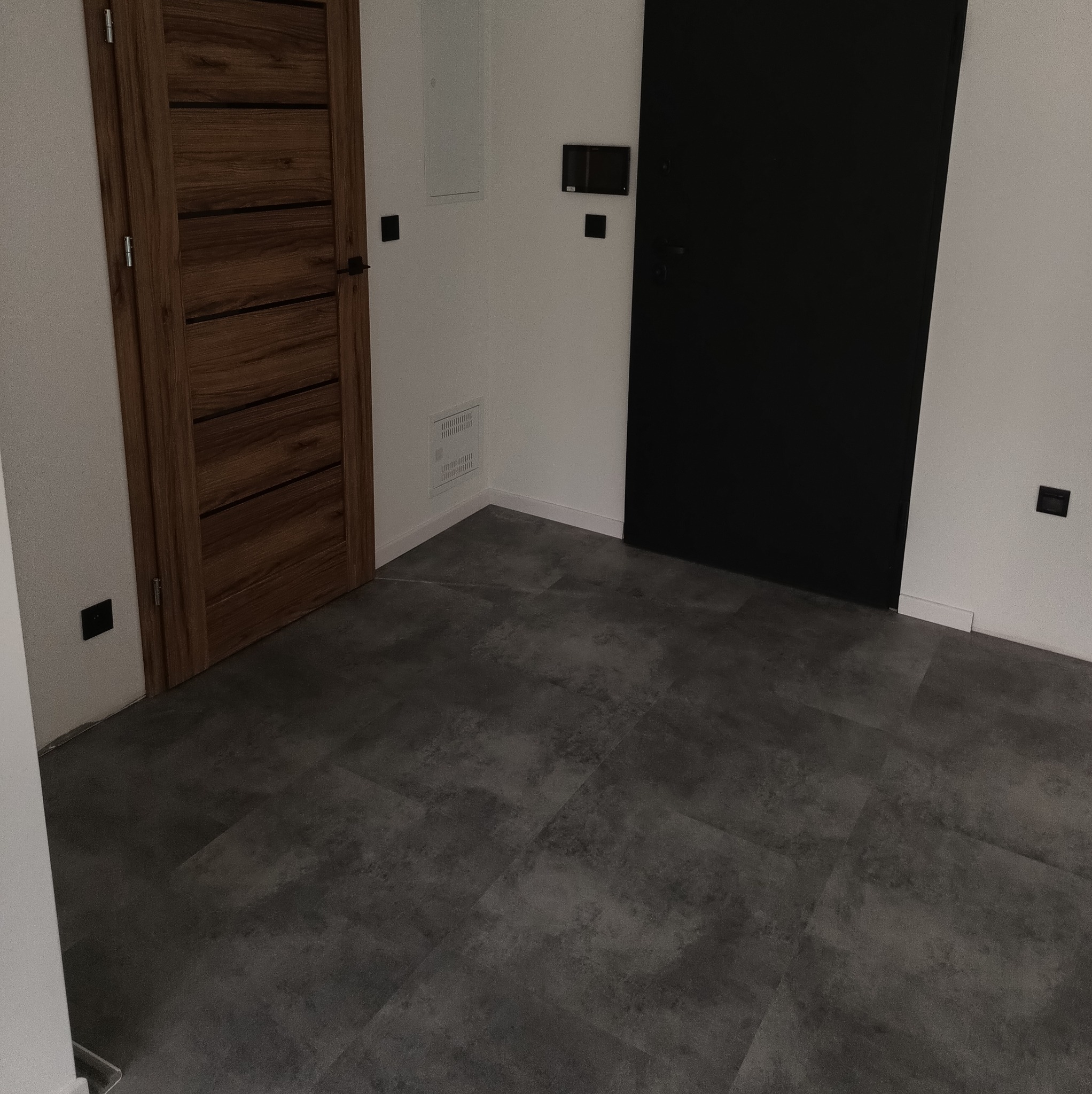 Nowoczesne wnętrze: szara podłoga z płytek, drewniane drzwi z poziomymi panelami i czarne drzwi w minimalistycznym stylu. Kontrastujące kolory i proste linie.