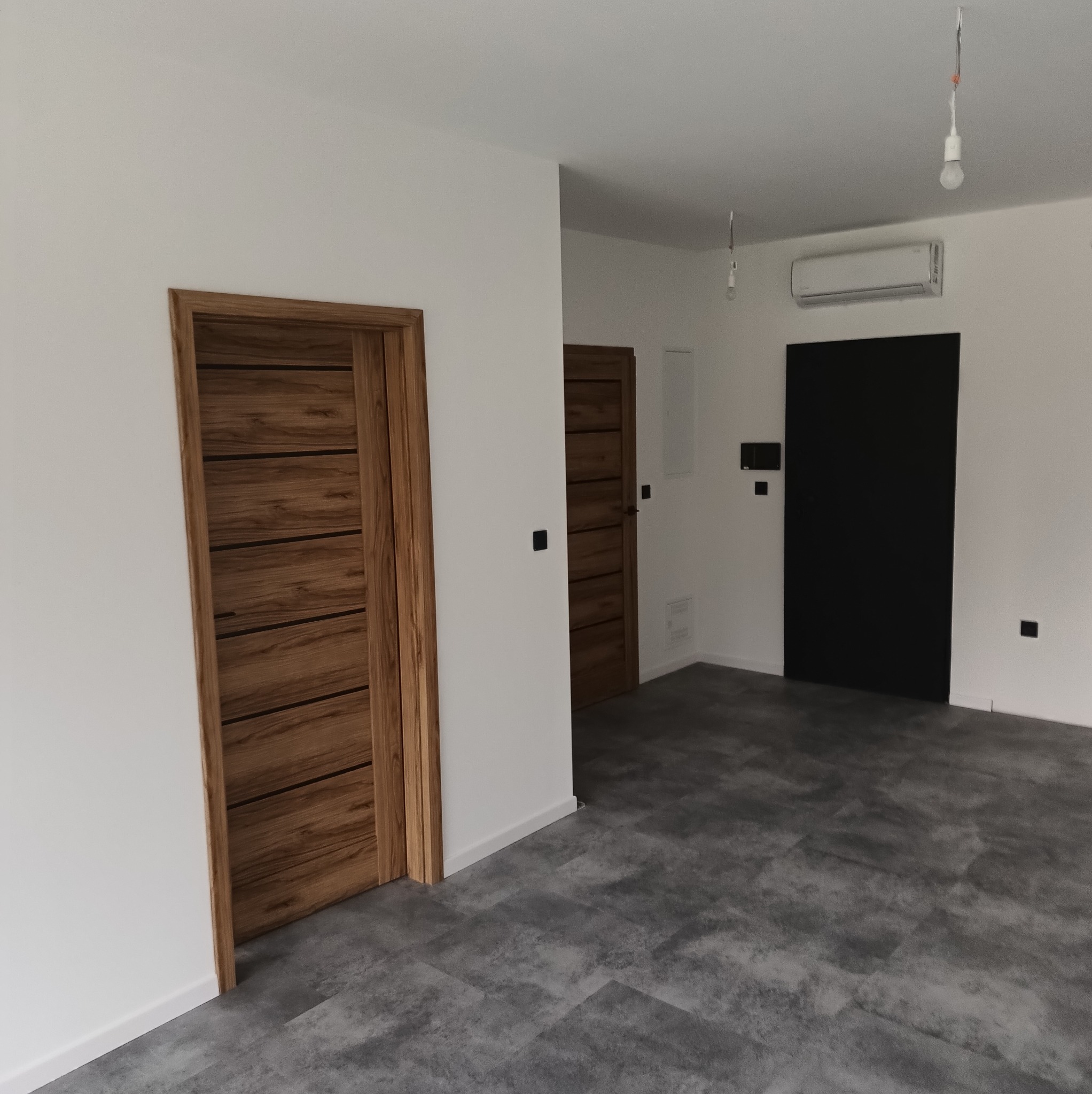 Nowoczesne wnętrze po remoncie w Mrągowie: drewniane drzwi z poziomymi panelami, szara podłoga imitująca beton, białe ściany i czarne drzwi w głębi.