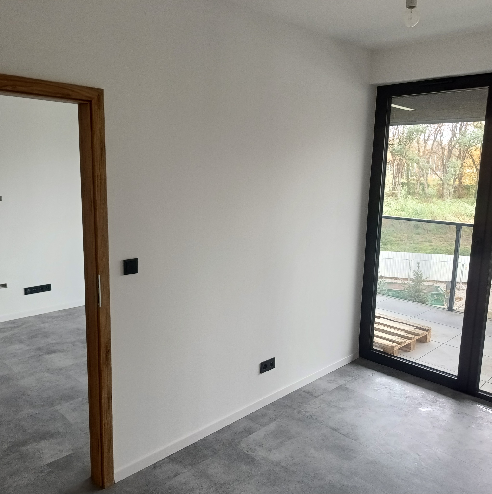 Wnętrze po odświeżeniu: białe ściany, szara podłoga imitująca beton, drewniana rama drzwi i duże okno balkonowe z widokiem na zieleń. Minimalistyczny design.