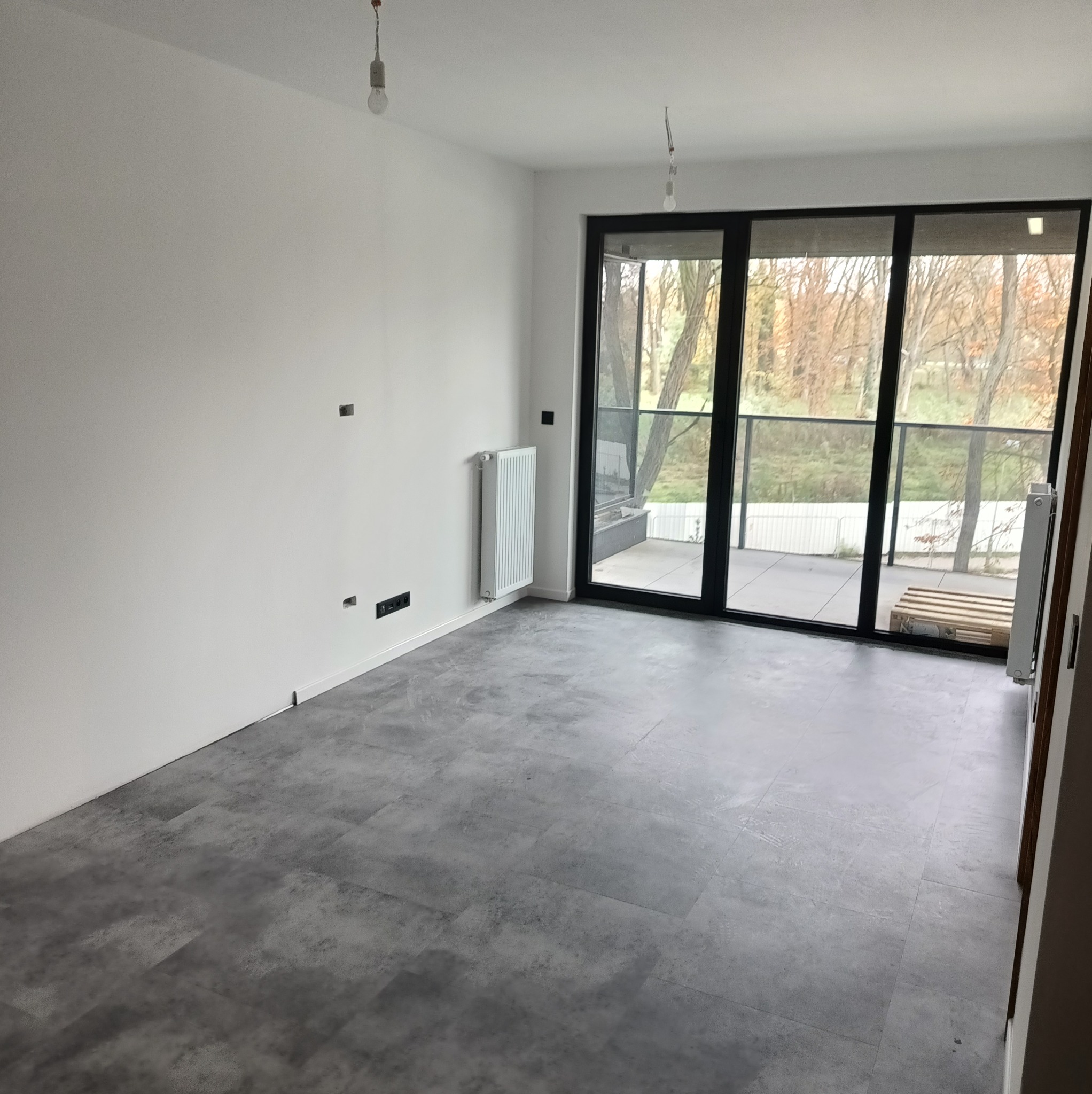 Wykończone wnętrze mieszkania z szarą podłogą, białymi ścianami, grzejnikiem i dużym oknem balkonowym z widokiem na drzewa. Widoczne gniazdka elektryczne.