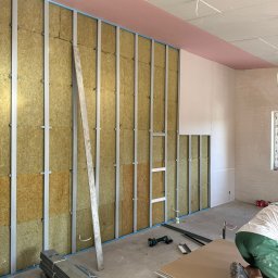 B.K. PROBUD SPÓŁKA Z OGRANICZONĄ ODPOWIEDZIALNOŚCIĄ - Montaż izolacji z wełny mineralnej w metalowym stelażu, częściowo zakrytej płytami gipsowo-kartonowymi. Widoczne narzędzia i materiały budowlane.