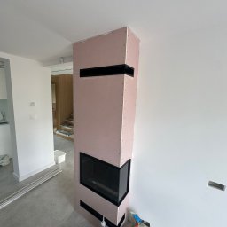 B.K. PROBUD SPÓŁKA Z OGRANICZONĄ ODPOWIEDZIALNOŚCIĄ - Obudowa kominka z płyty GK w trakcie montażu, widoczne czarne wnęki wentylacyjne. Ściana pomalowana na biało, na podłodze szare płytki. W tle widoczne schody.