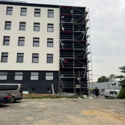 B.K. PROBUD SPÓŁKA Z OGRANICZONĄ ODPOWIEDZIALNOŚCIĄ - Budynek w trakcie prac elewacyjnych z rusztowaniem. Część fasady biała, część czarna. Widoczni pracownicy i zaparkowane samochody. Pochmurne niebo w tle.