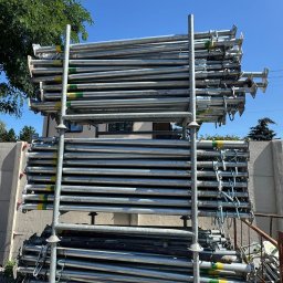 B.K. PROBUD SPÓŁKA Z OGRANICZONĄ ODPOWIEDZIALNOŚCIĄ - Stalowe stemple budowlane ułożone na regale, widoczne zielono-żółte oznaczenia, w tle budynek i błękitne niebo. Sprzęt budowlany gotowy do użycia.