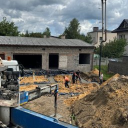B.K. PROBUD SPÓŁKA Z OGRANICZONĄ ODPOWIEDZIALNOŚCIĄ - Fundamenty budynku w trakcie izolacji. Widoczny sprzęt budowlany, piasek, folia izolacyjna i pracownicy. Pochmurne niebo w tle.