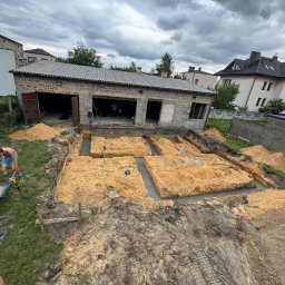 B.K. PROBUD SPÓŁKA Z OGRANICZONĄ ODPOWIEDZIALNOŚCIĄ - Wykopane fundamenty pod budynek z częściowo wylanym betonem, widoczne narzędzia i pracownik z taczką. Prace ziemne w toku, pochmurne niebo w tle.