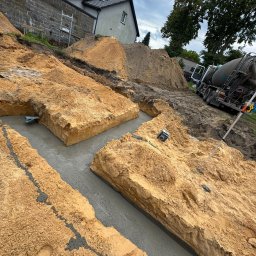 B.K. PROBUD SPÓŁKA Z OGRANICZONĄ ODPOWIEDZIALNOŚCIĄ - Wykopane fundamenty zalane betonem, widoczny mikser do betonu w tle. Prace ziemne i betonowe w trakcie realizacji. Ujęcie z góry, pochmurne niebo.