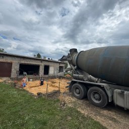 B.K. PROBUD SPÓŁKA Z OGRANICZONĄ ODPOWIEDZIALNOŚCIĄ - Betoniarka na budowie domu jednorodzinnego. Wykopane fundamenty, widoczni pracownicy. Pochmurne niebo. Etap wylewania fundamentów.