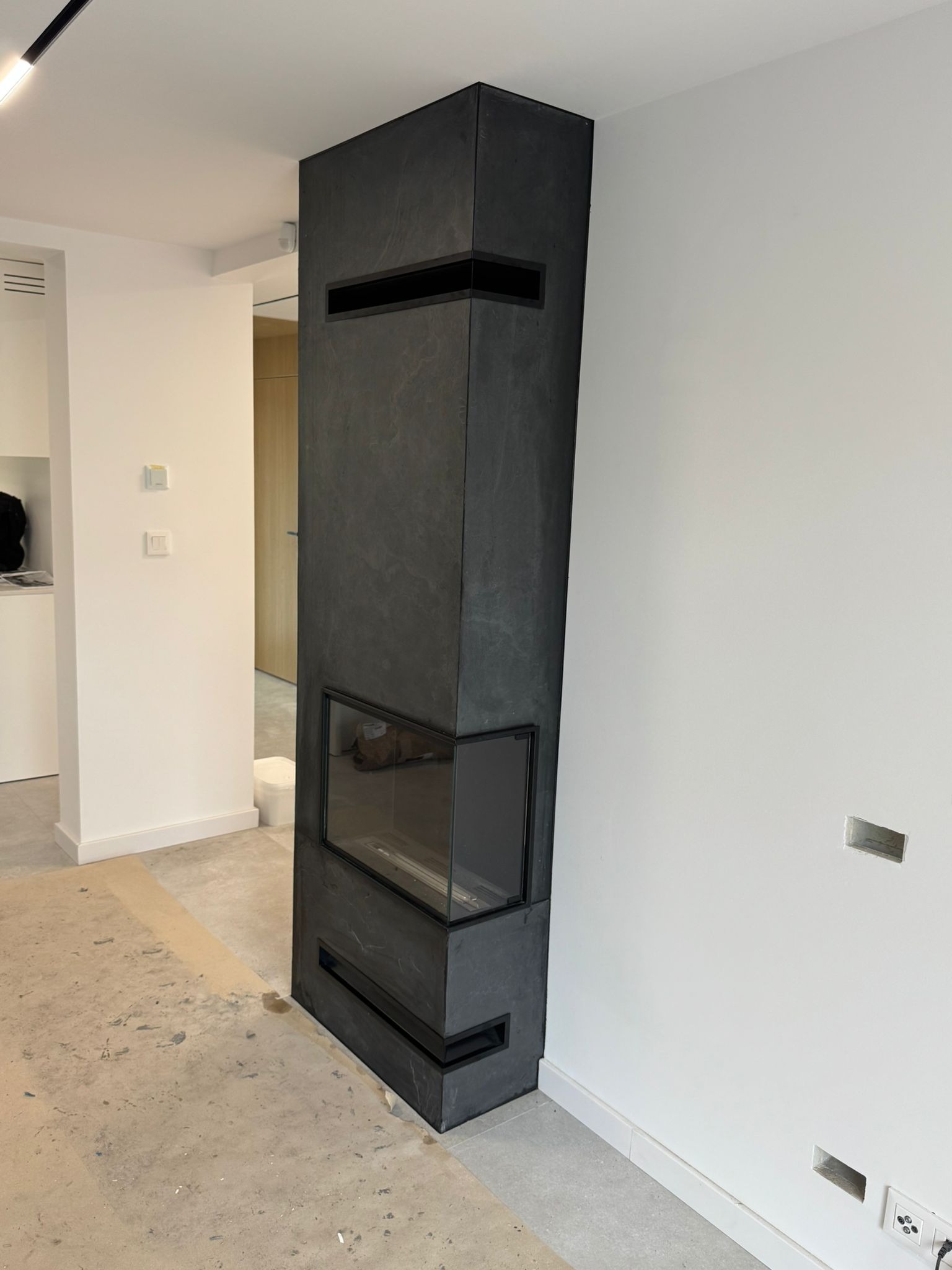 Nowoczesny kominek obudowany ciemnym kamieniem, z widocznym paleniskiem i elementami dekoracyjnymi, w minimalistycznym wnętrzu z białymi ścianami i surową podłogą.