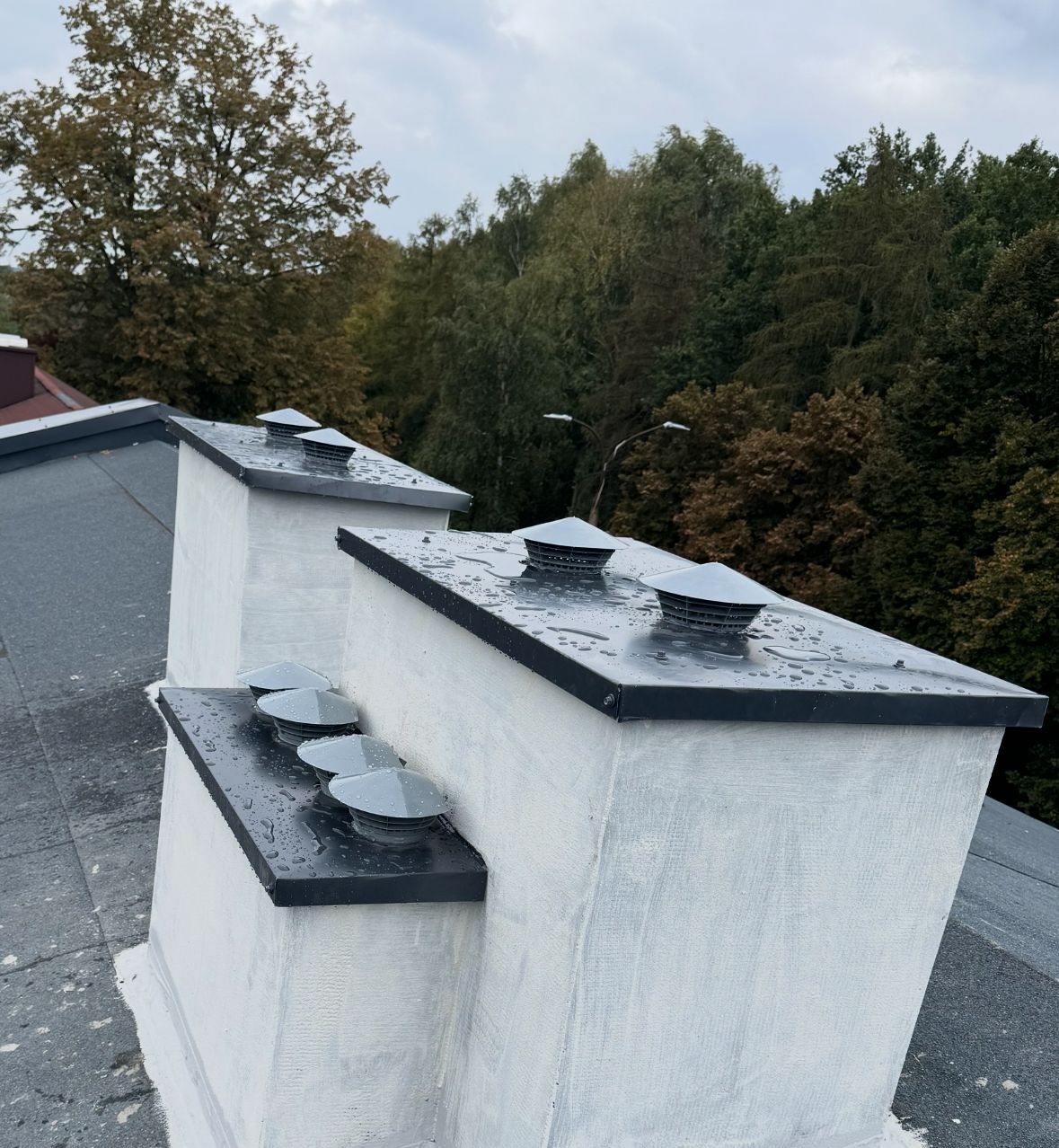 Otynkowane kominy z metalowymi daszkami na dachu pokrytym papą, widoczne krople deszczu. W tle drzewa i pochmurne niebo. Ujęcie z dołu, podkreślające detale wykończenia.
