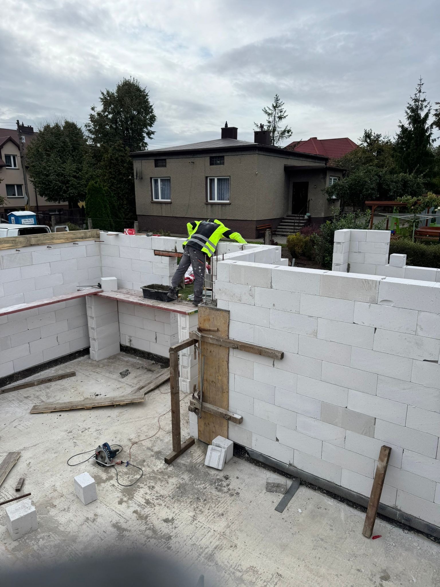 Budowa domu z białych bloczków: murarz w żółtej kamizelce układa ściany. Widoczne narzędzia i elementy konstrukcyjne. W tle domy i pochmurne niebo.