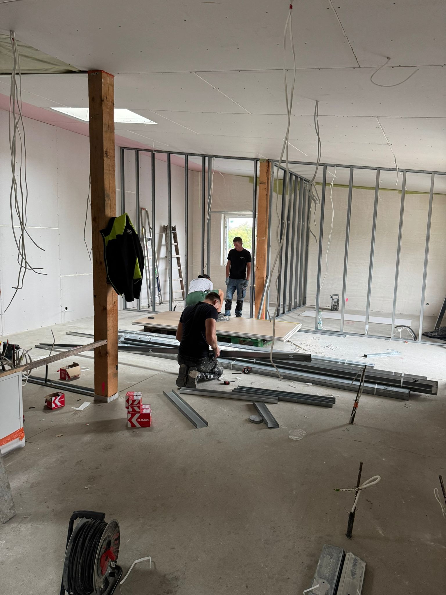 Montaż ścian działowych z płyt gipsowo-kartonowych w surowym wnętrzu. Widoczne profile, płyty, narzędzia i pracownicy w trakcie pracy. Oświetlenie sufitowe.