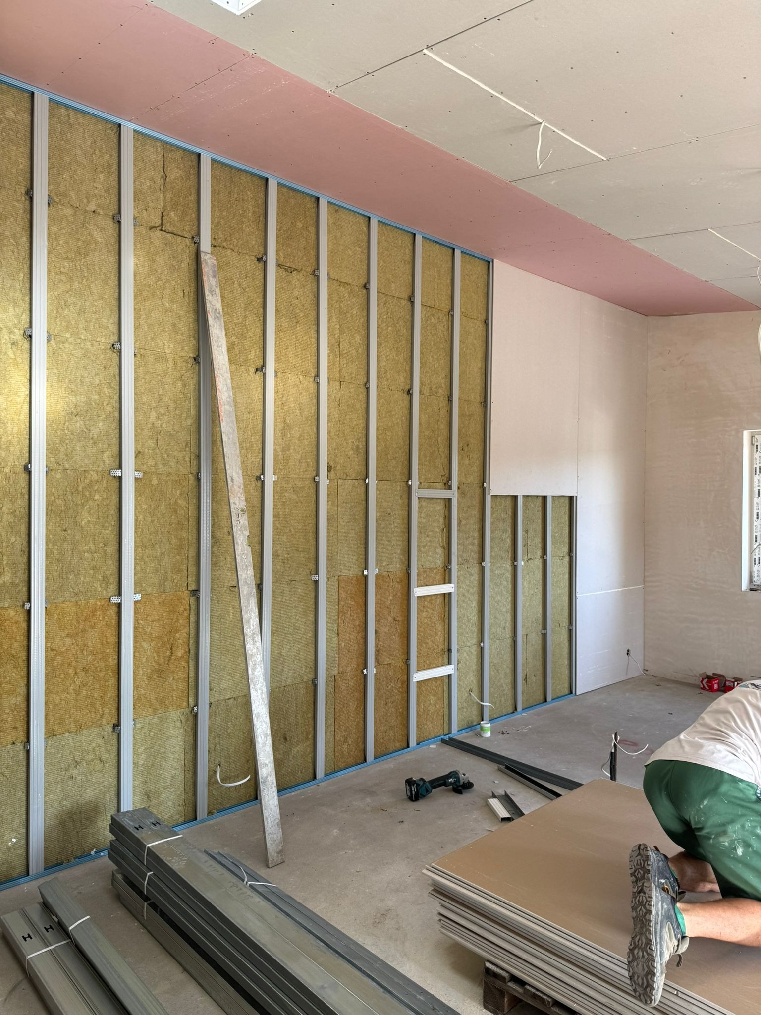 Montaż izolacji z wełny mineralnej w metalowym stelażu, częściowo zakrytej płytami gipsowo-kartonowymi. Widoczne narzędzia i materiały budowlane.