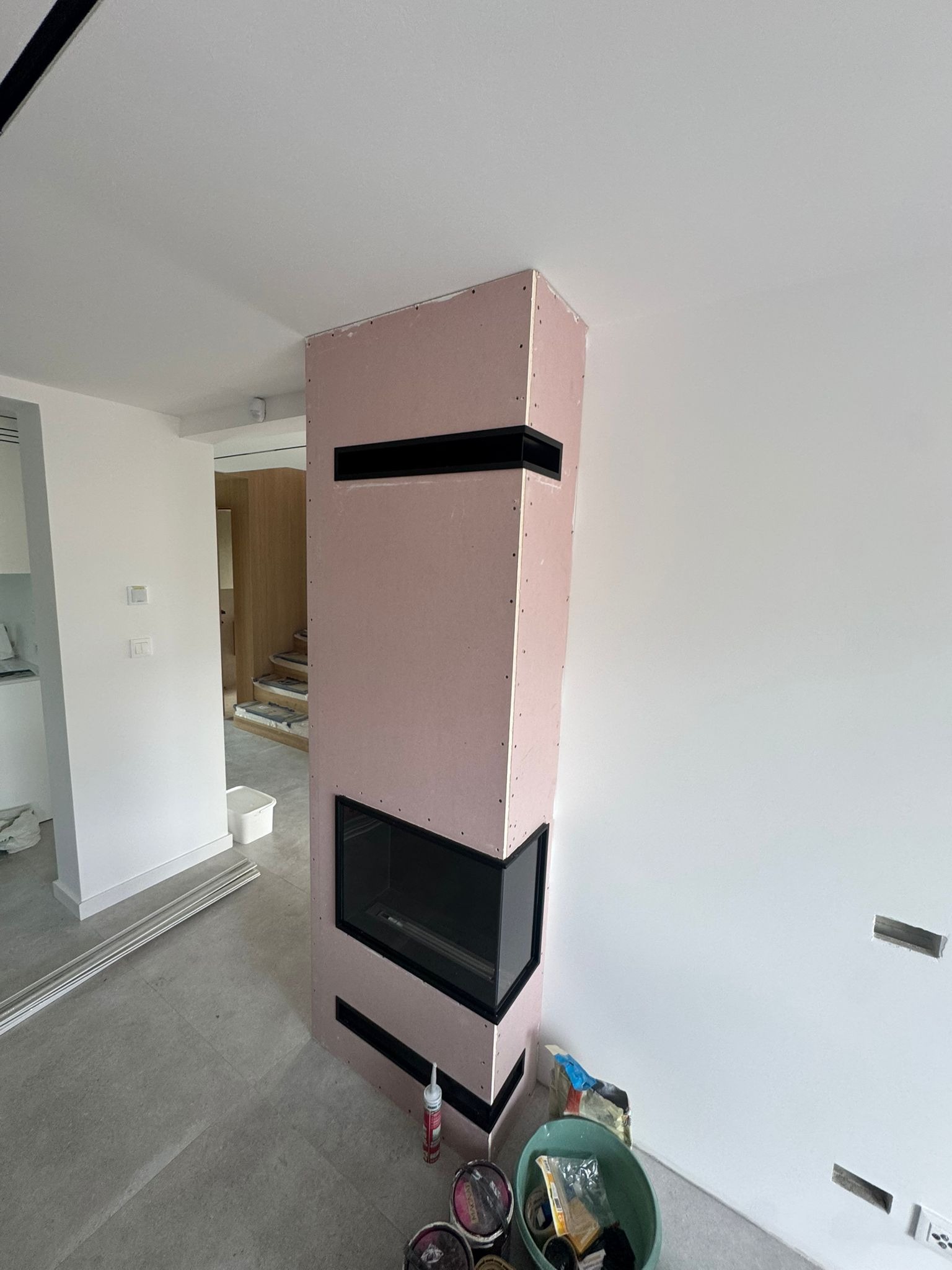 Obudowa kominka z płyty GK w trakcie montażu, widoczne czarne wnęki wentylacyjne. Ściana pomalowana na biało, na podłodze szare płytki. W tle widoczne schody.