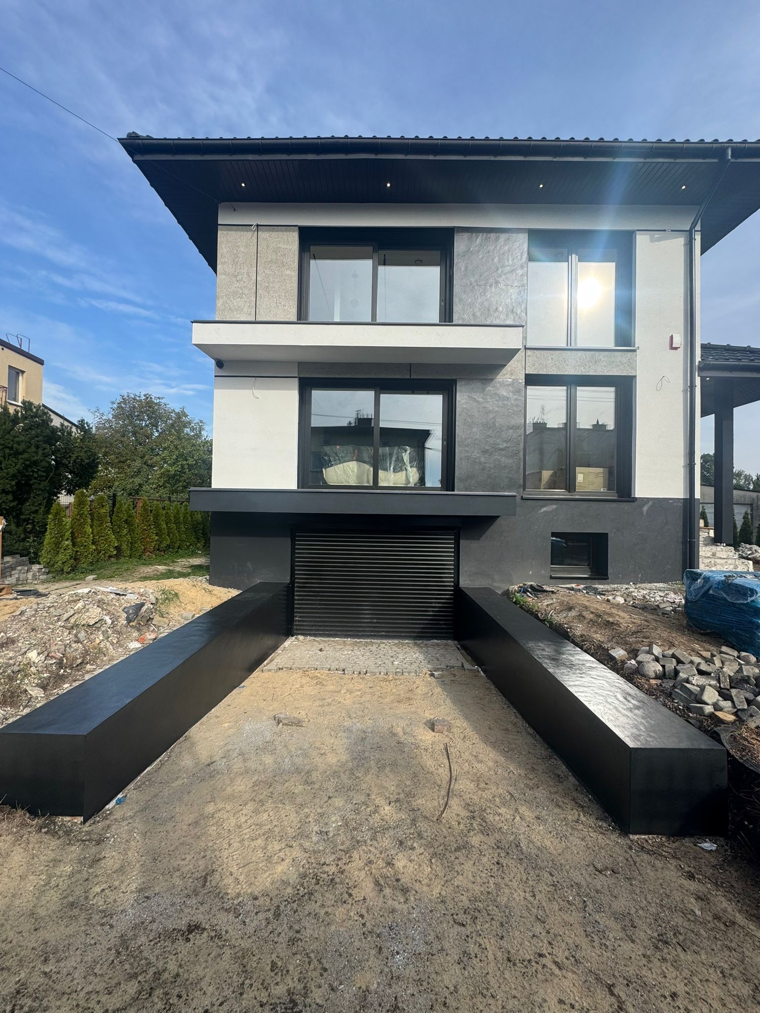 Dwukondygnacyjny dom z garażem w piwnicy, wykończony w odcieniach szarości i bieli, z widocznymi elementami elewacji i zagospodarowania terenu.